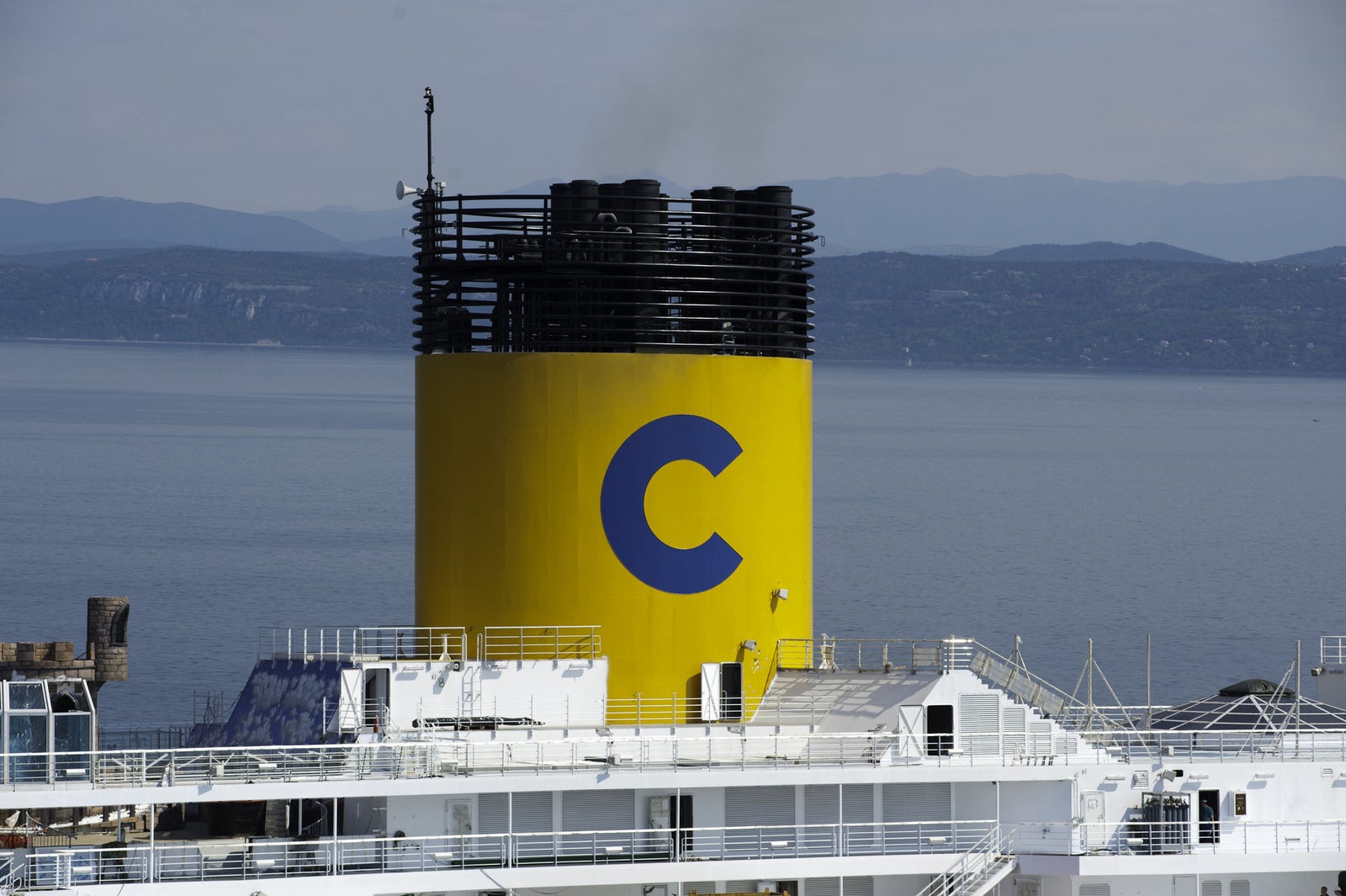 Fincantieri | Group - Business - Products - Cruise Ships - Costa Favolosa: Close-up view of ship’s bright yellow chimney with large blue 'C' logo, vessel docked near calm waterfront, distant mountains in background, industrial design contrasts with serene natural setting.