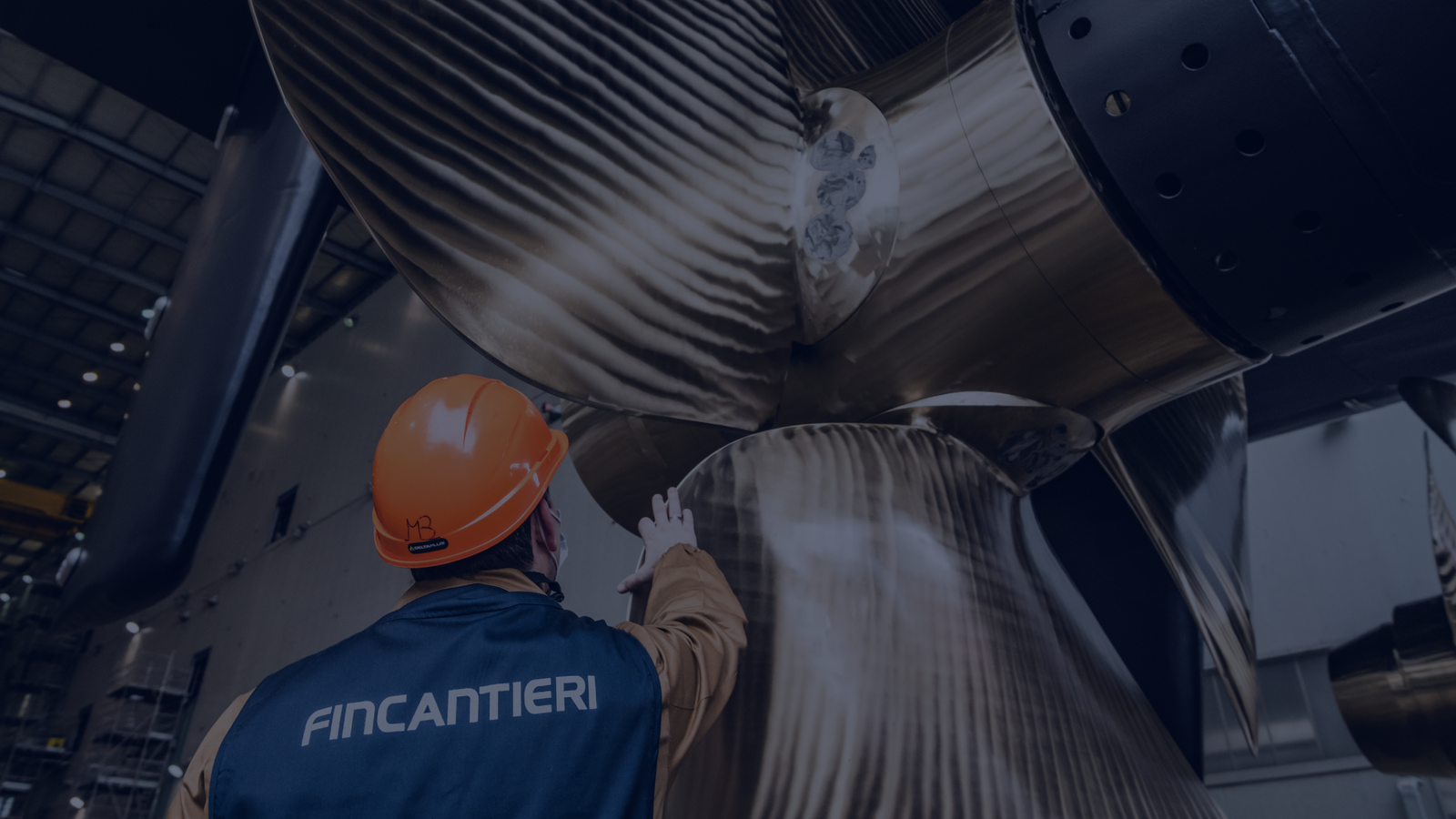 Fincantieri - People: Worker wearing orange safety helmet and Fincantieri jacket inspects large metallic ship propeller in shipyard or industrial facility, structural details in the background, propeller with textured and polished finish.