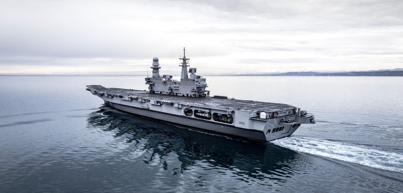 Fincantieri | Group - Business - Products - Naval Vessels - Aircraft Carriers and Amphibious Ships - Cavour: Rear lateral view of aircraft carrier "Cavour" in operation, gray hull, long flight deck, prominent island structure, sailing on open water, cloudy sky.