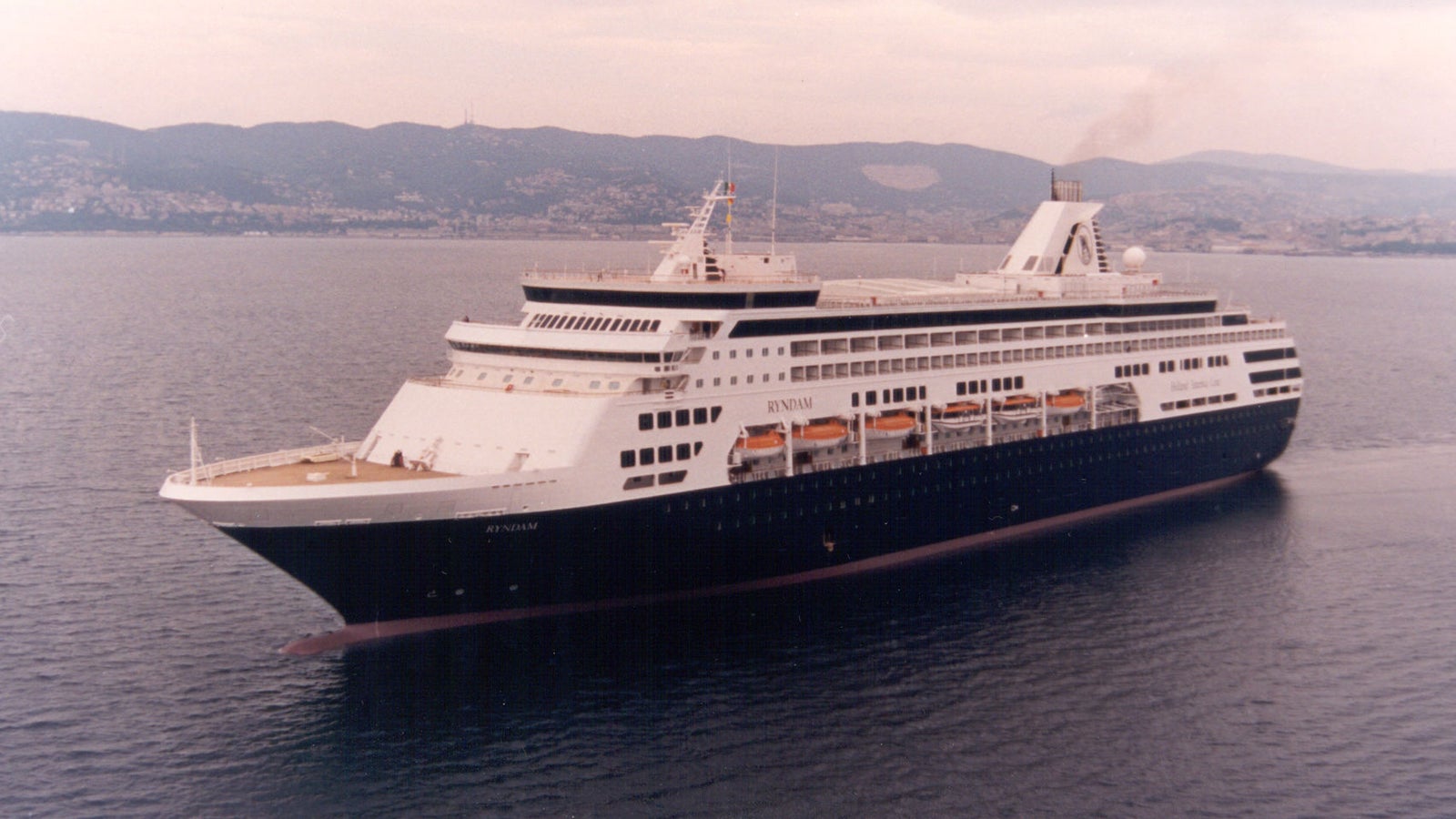 Fincantieri | Group - Business - Products - Cruise Ships - Ryndam: Side view of Ryndam cruise ship in operation navigating, multiple decks and visible lifeboats, mountainous coastal background, overcast sky.