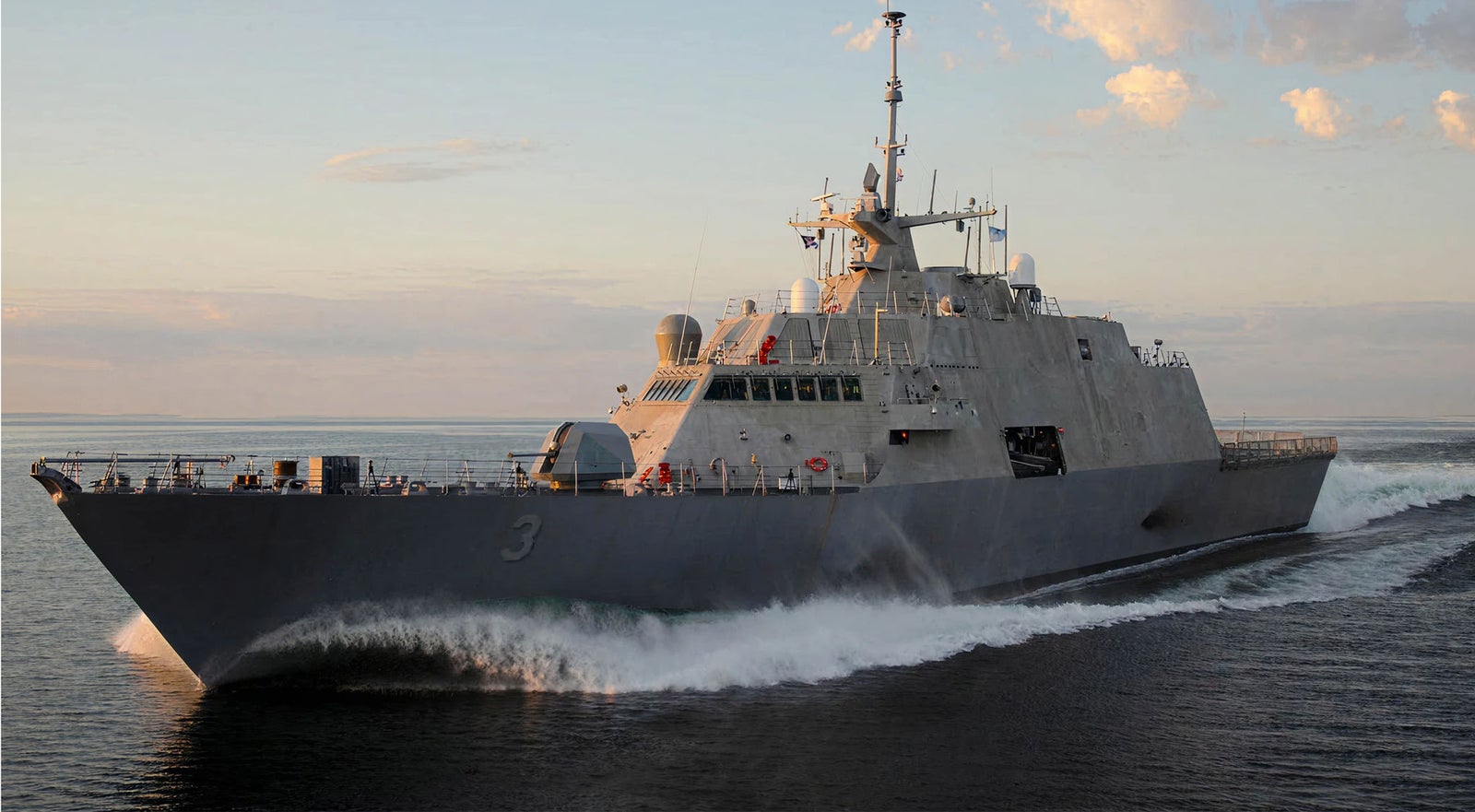 Fincantieri | Gruppo - Società - Controllate e collegate - Marine Systems North America: Grande nave militare operativa in navigazione su acque aperte al tramonto, design moderno e filante, antenne ed equipaggiamenti visibili sul ponte; numero ‘3’ ben visibile sullo scafo, onde calme e cielo soft con nuvole sparse che valorizzano la scena.