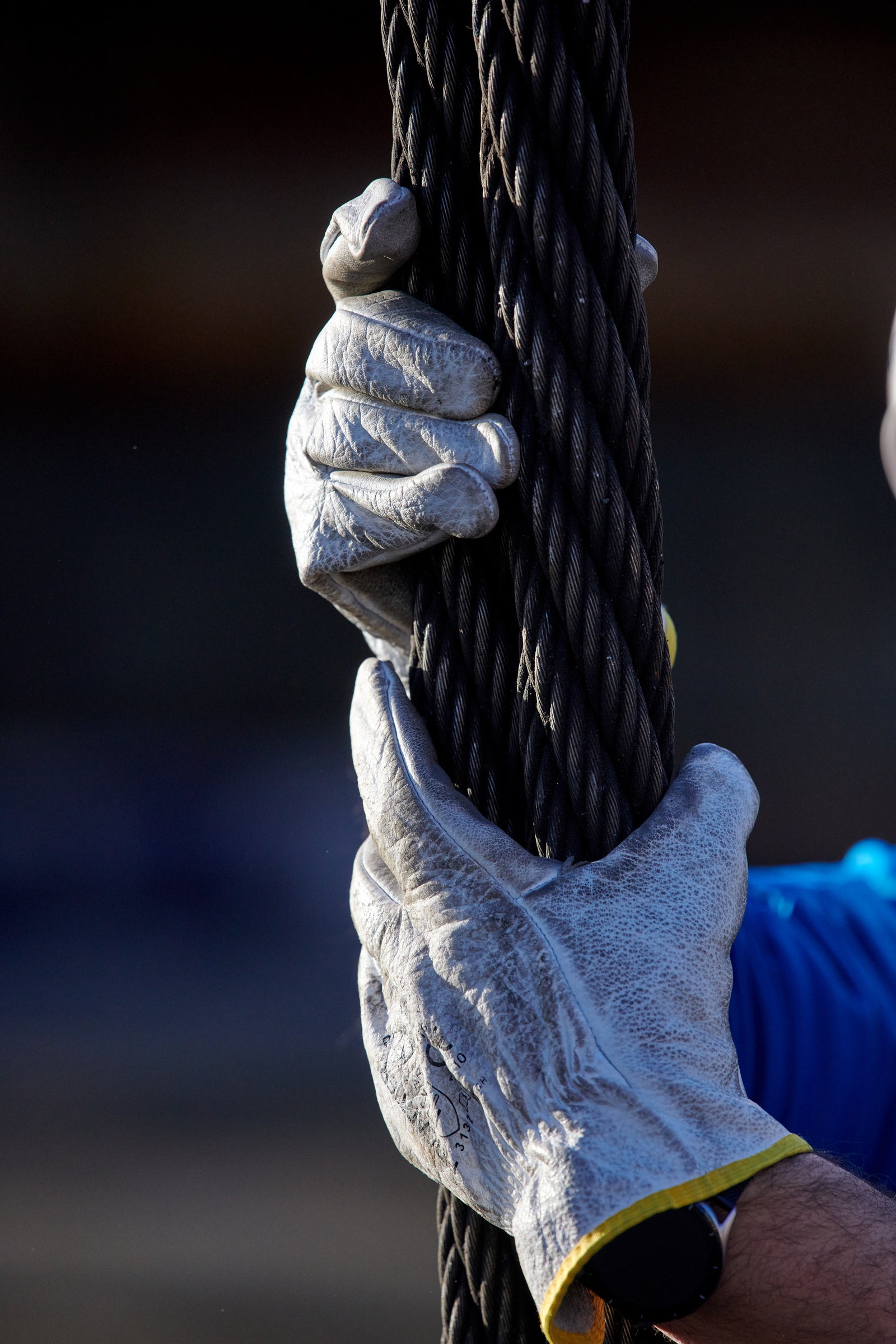 Fincantieri | Group - Sustainability - Industrial Excellence - Health and Safety: Close-up of gloved hands firmly holding a thick steel cable, gloves with visible stitching and reinforced materials, blurred background. Industrial or shipyard work context.
