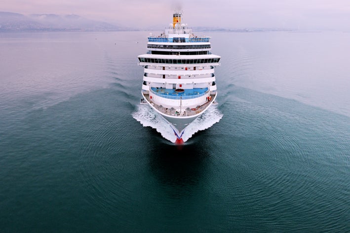  Fincantieri | Group - Business - Products - Cruise Ships - Costa Deliziosa: Large white cruise ship in operation sailing through calm open waters, multiple decks, distinctive red and blue design at bow, serene setting with soft overcast sky and faint distant mountains, gentle ripples as ship moves forward.