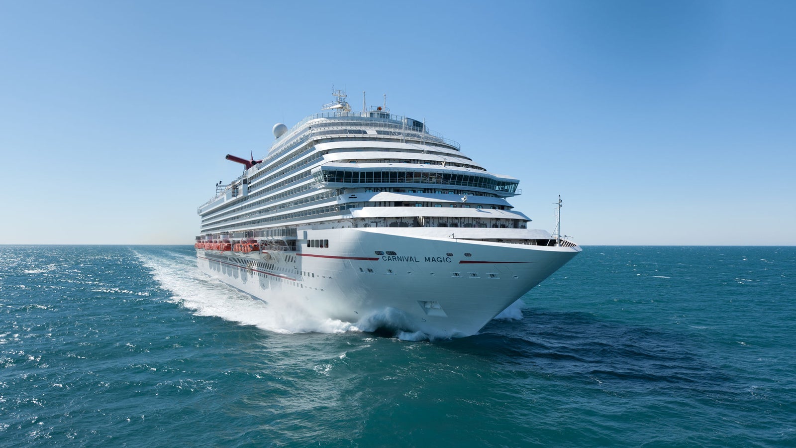 Fincantieri | Group - Business - Products - Cruise Ships - Carnival Magic: Side view of Carnival Magic cruise ship in operation navigating calm open waters under clear blue sky, name visible on hull, multi-deck structure sunlit, scene captures ship’s grandeur and serene ocean environment.