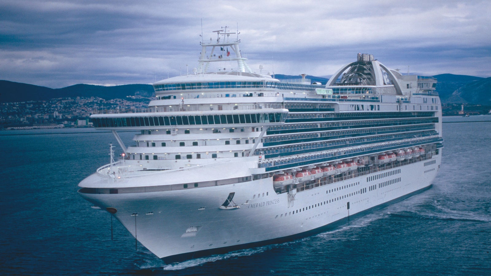 Fincantieri | Group - Business - Products - Cruise Ships - Emerald Princess: Side view of Emerald Princess cruise ship in operation navigating calm waters near coastline, white hull, multiple passenger decks, visible balconies, coastal city and mountains in background.