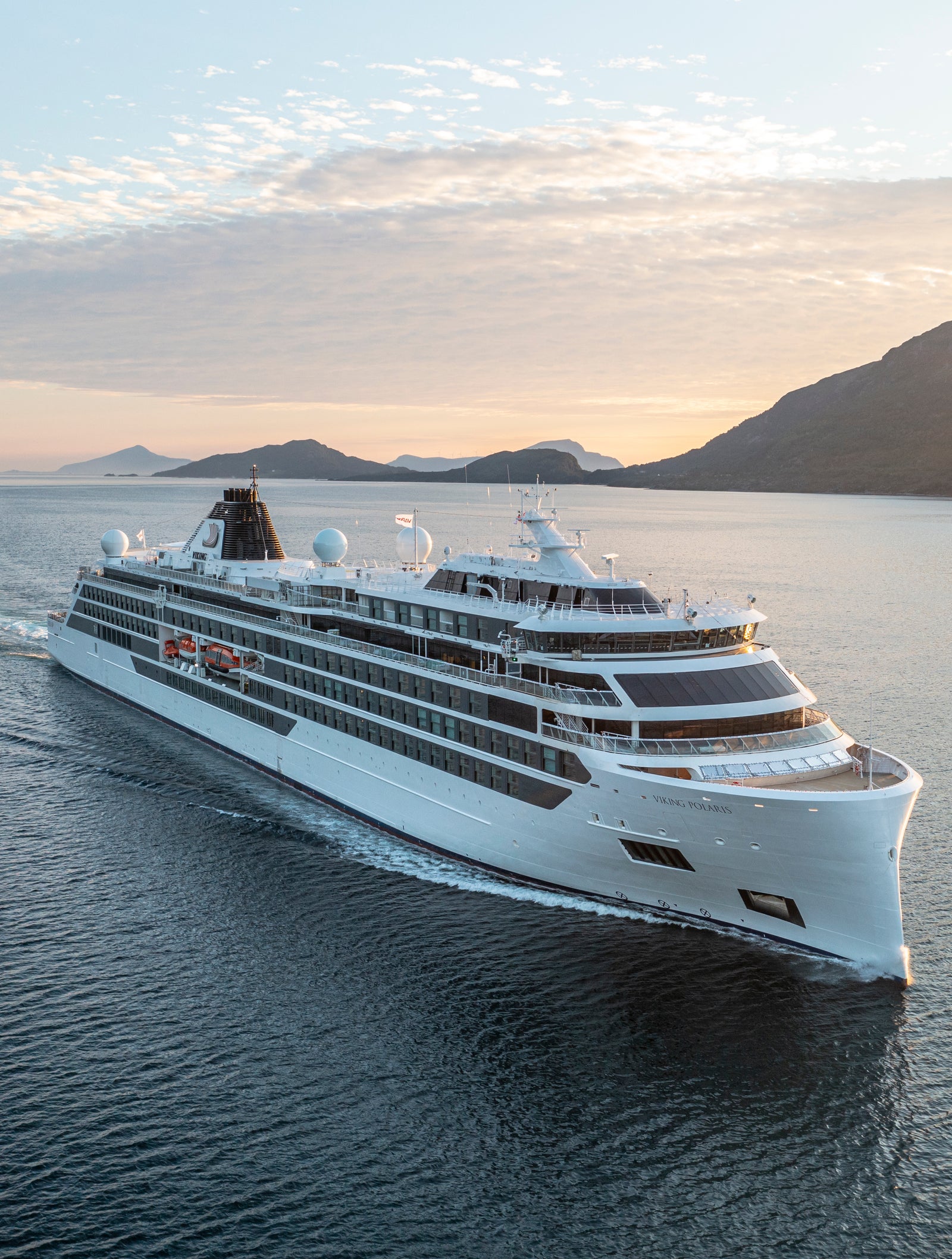 Fincantieri | Group - Business - Products - Cruise Ships - Viking Polaris: Modern luxury cruise ship in operation, gliding through calm waters at sunrise, sleek white design with visible balconies, scenic mountains and serene sky emphasizing grandeur and travel.