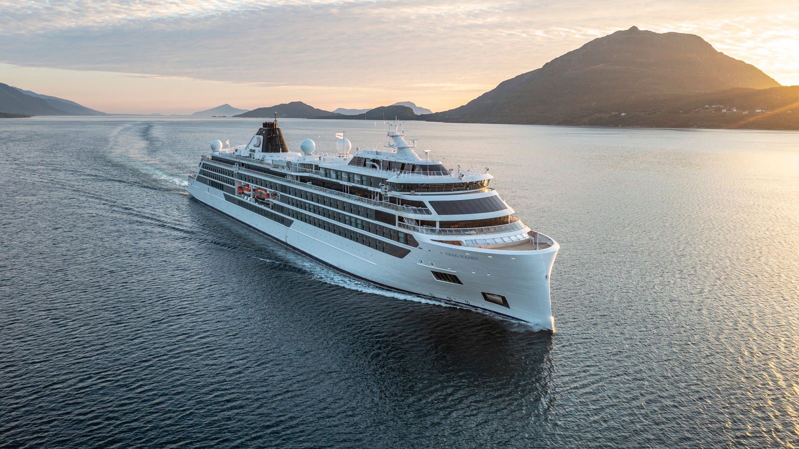 Fincantieri | Group - Business - Products - Cruise Ships - Viking Polaris: Side view of Viking Polaris cruise ship in operation, modern white design with visible balconies, navigating at sunrise, mountainous background.
