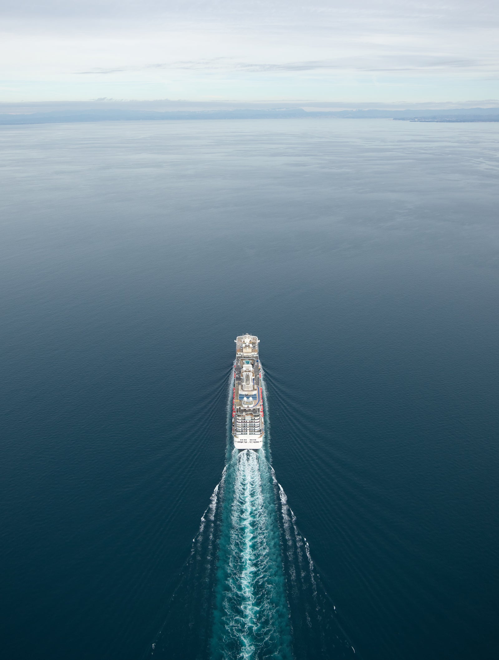 Fincantieri | Group - Business - Products - Cruise Ships - Discovery Princess: Overhead stern view of Discovery Princess cruise ship in operation, visible wake trailing into blue sea, calm and serene marine environment.