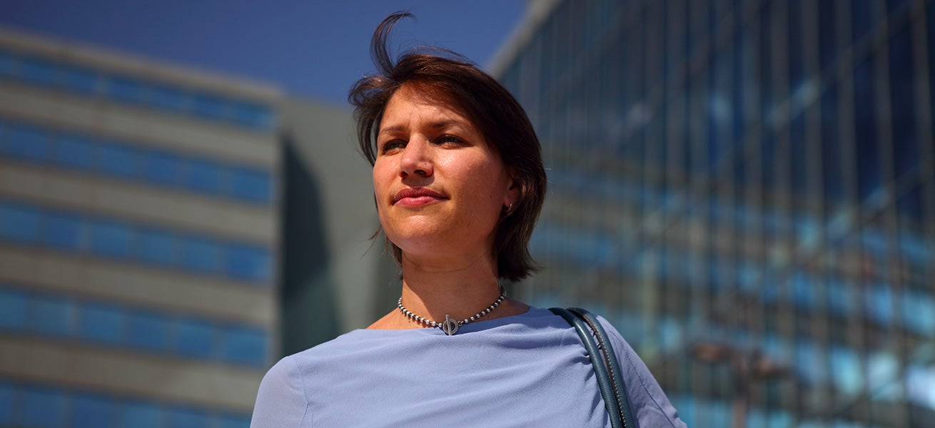 Fincantieri – People: Woman wearing a light blue top and pearl necklace in an outdoor setting in front of modern glass buildings, blue sky and contemporary architecture, professional or informal urban atmosphere.