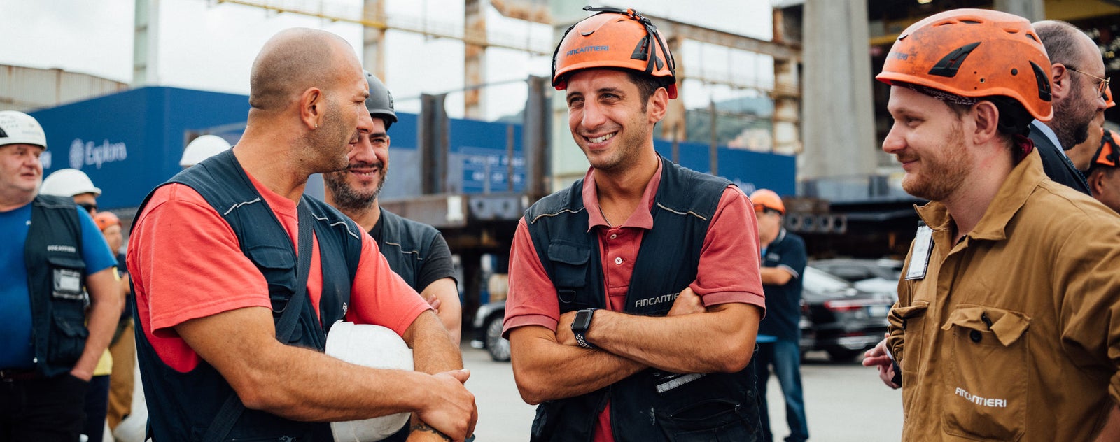 Fincantieri | Sostenibilità - Eccellenza industriale - Squadra operai: Gruppo di lavoratori con caschi e giubbotti di sicurezza riuniti in cantiere industriale, con strutture e ponteggi sullo sfondo. Discussione focalizzata su lavoro di squadra e sicurezza; testo 'Epiroc' visibile su edificio.