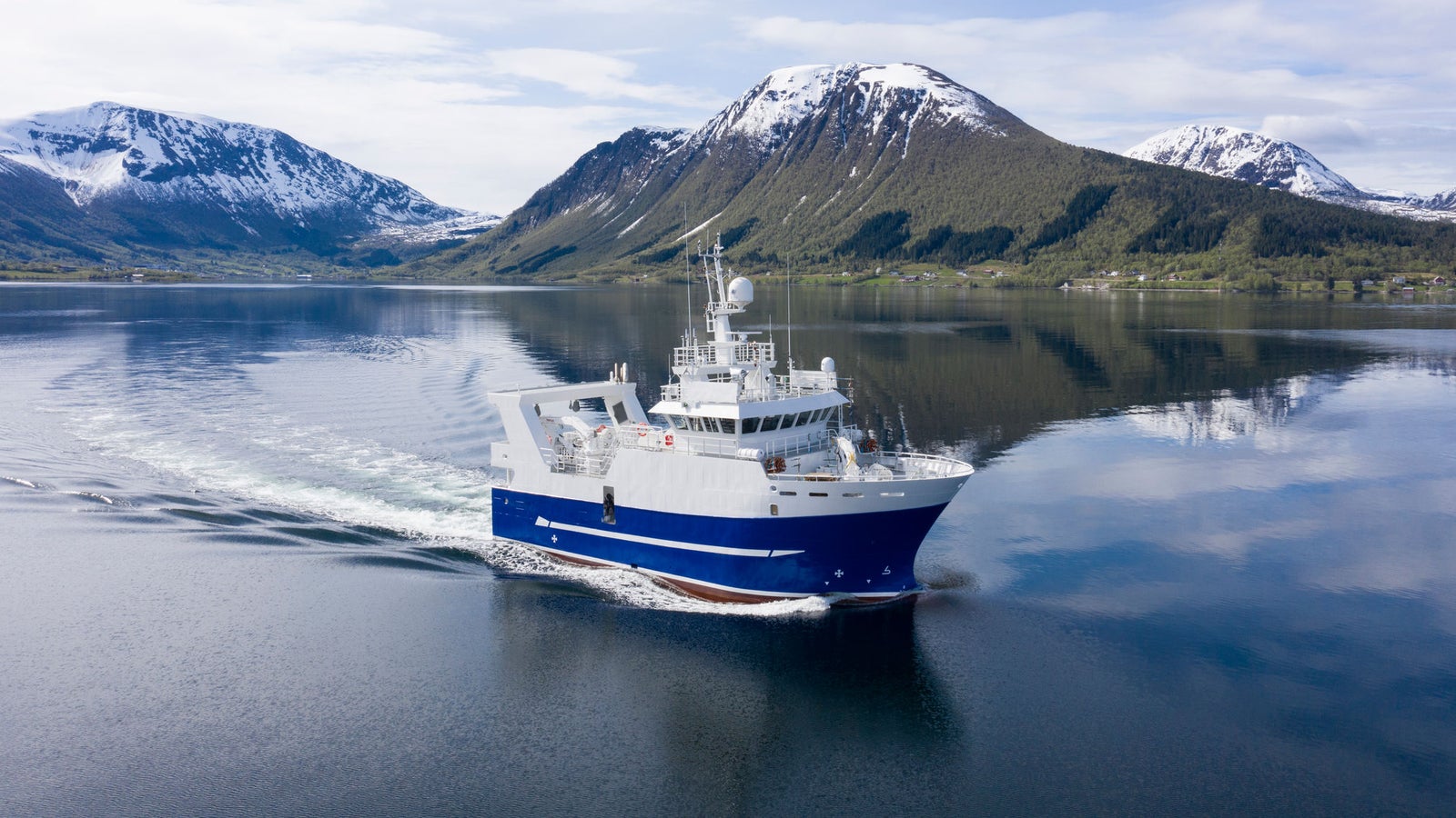 Prodotto - Peschereccio Remoybuen: Vista laterale della nave operativa in navigazione in un fiordo, scafo blu e bianco, scia visibile, sfondo con montagne innevate, design moderno.