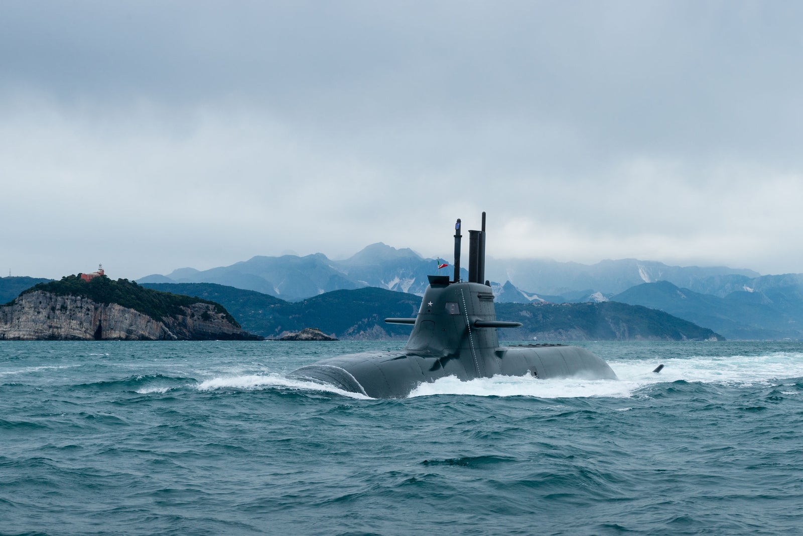 Fincantieri | Chi siamo - Eredità e Futuro: Sottomarino militare dal design slanciato e funzionale, antenne e periscopi visibili, in navigazione parzialmente immerso su acque calme, sfondo costiero montuoso e cielo nuvoloso.