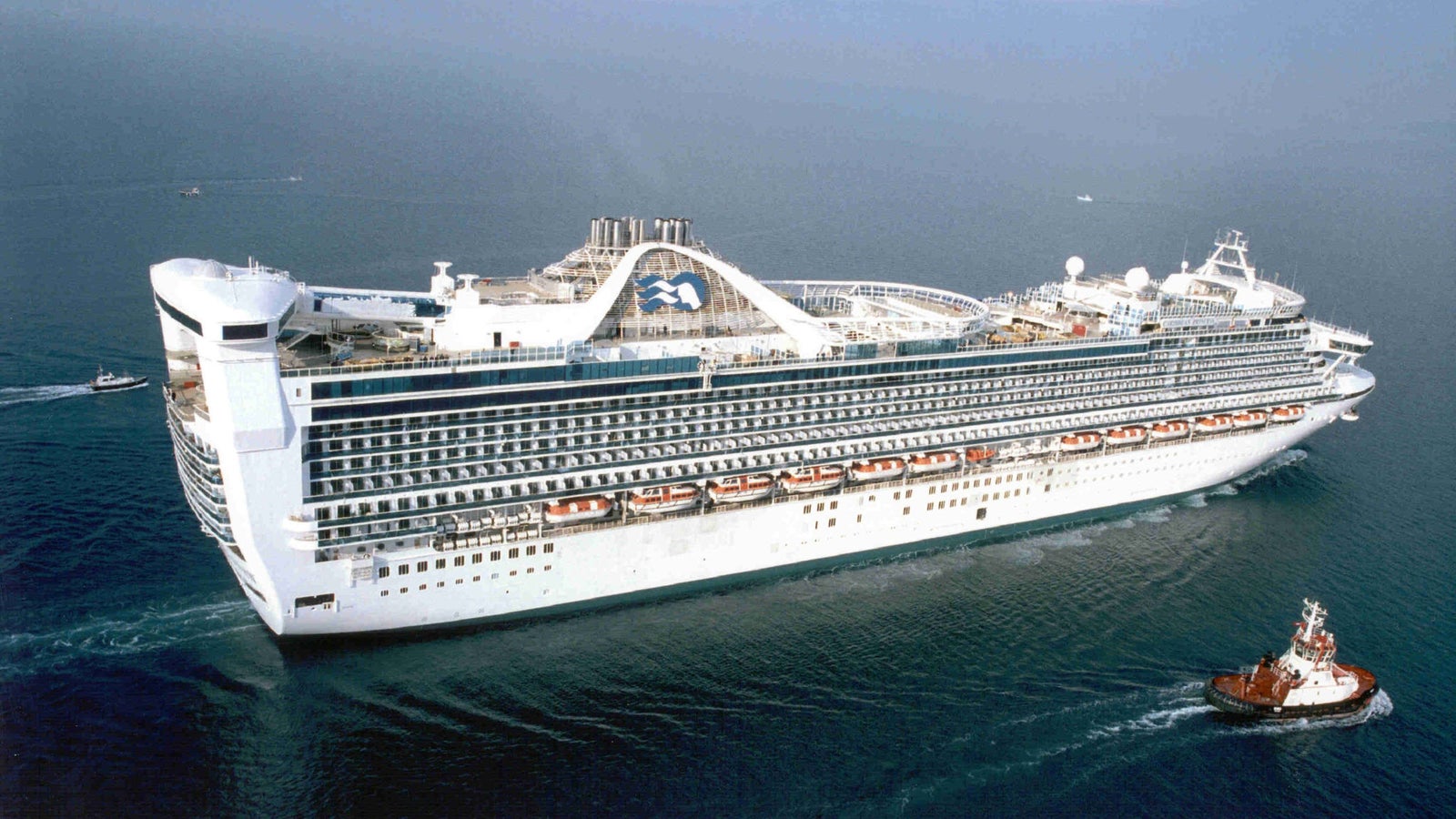 Fincantieri | Gruppo - Business - Prodotti - Navi da Crociera - Caribbean Princess: Vista laterale della nave Caribbean Princess operativa, scafo bianco, multipli ponti e balconi, logo a forma di corona visibile, trainata da rimorchiatore.