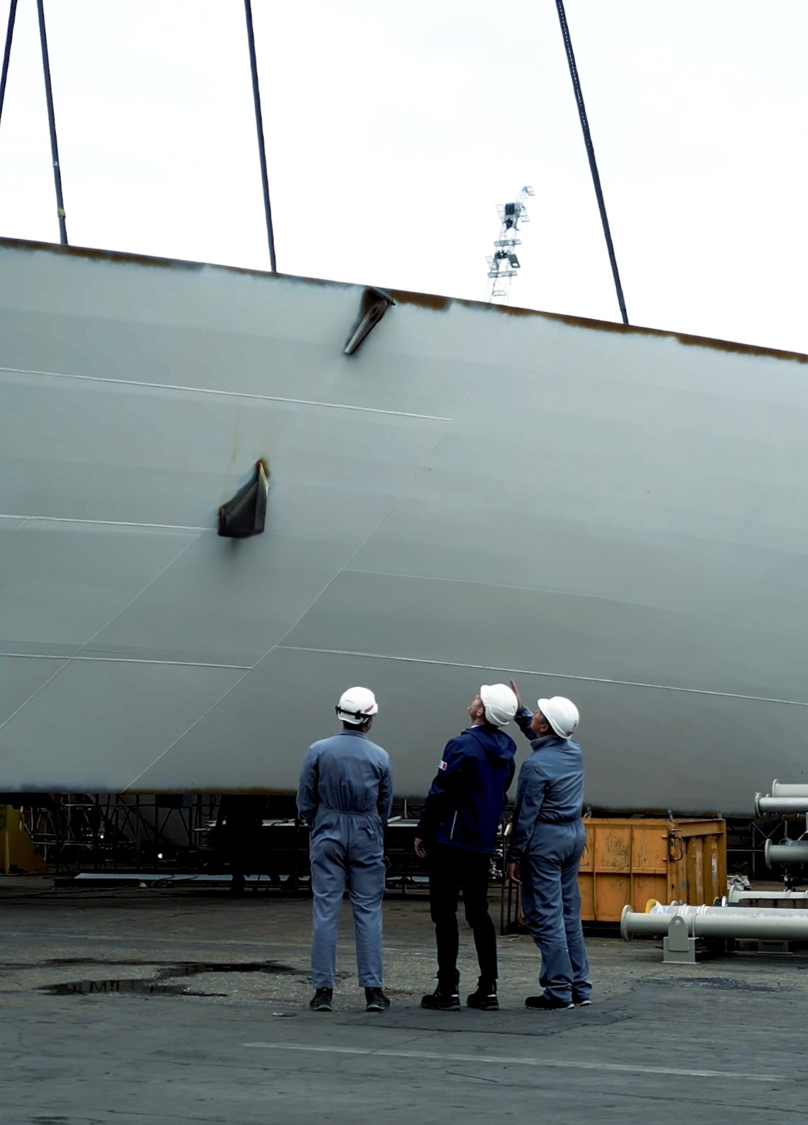 Fincantieri | Group - Sustainability - People and Community - Community: Four people, three wearing helmets and work uniforms, on an industrial pier examining the hull of a large grey ship; visible equipment and boxes, crane partially overhead the group.