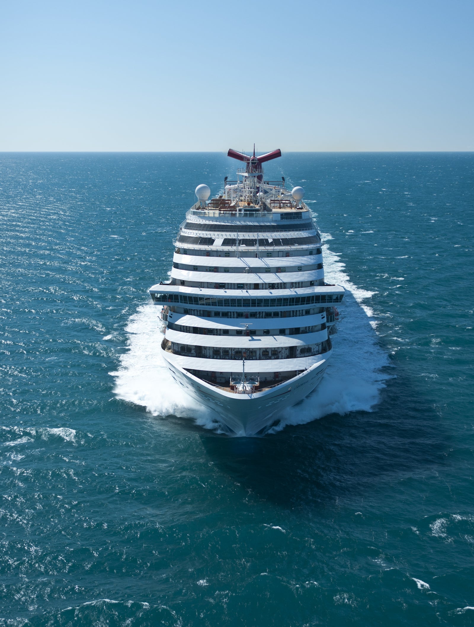 Fincantieri | Group - Business - Products - Cruise Ships - Carnival Magic: Massive cruise ship in operation sailing through open ocean under clear blue sky, multiple decks and distinctive red funnel, calm waters and bright sunlight create serene and adventurous atmosphere.