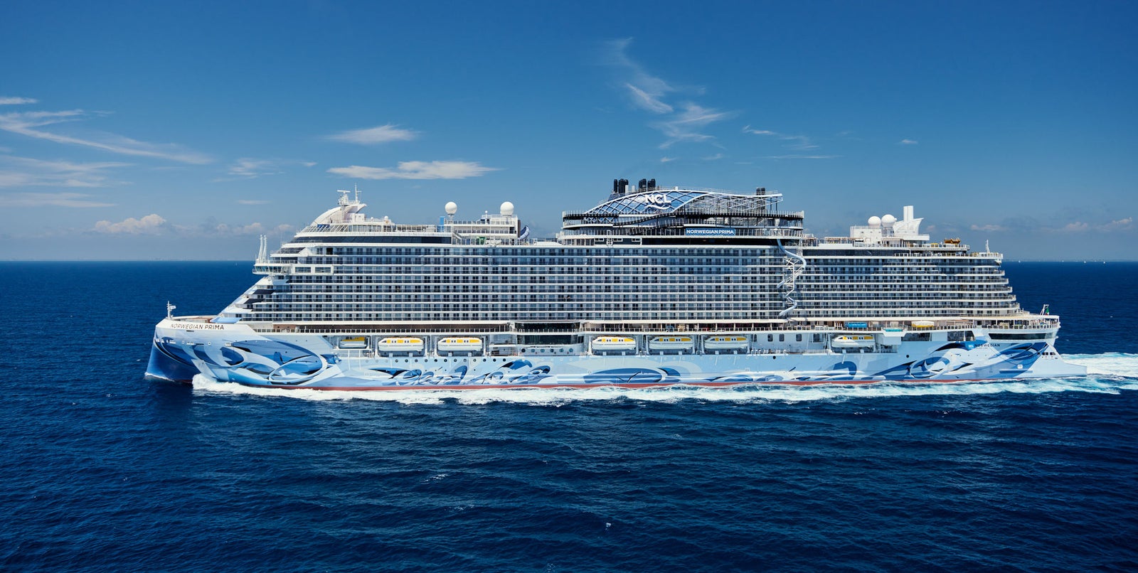 Fincantieri | Group - Business - Products - Cruise Ships - Norwegian Prima: Side view of Norwegian Prima cruise ship in operation, multiple decks and amenities, elaborate marine artwork on hull, large luxurious structure.