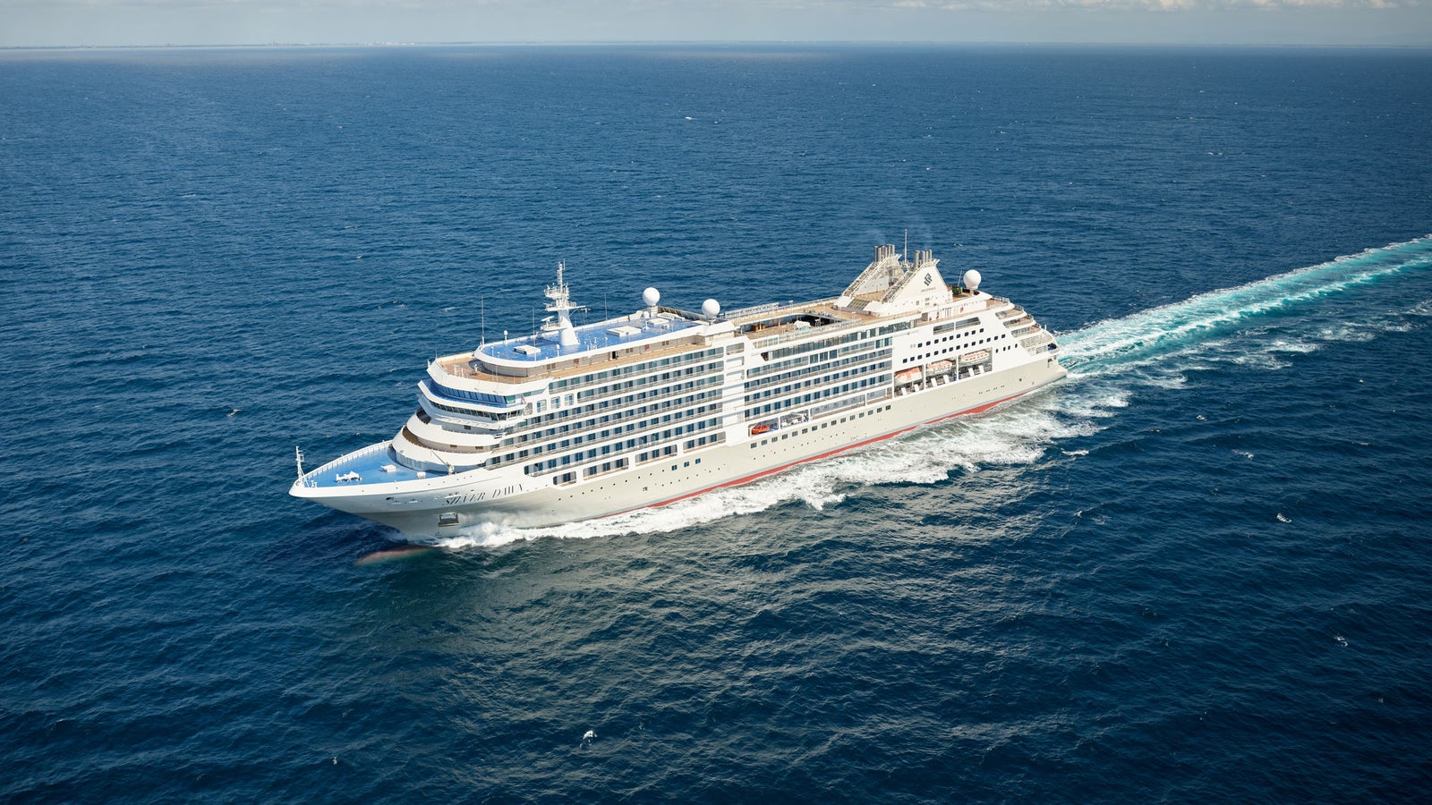 Fincantieri | Group - Business - Products - Cruise Ships - Silver Dawn: Large modern cruise ship in operation gliding through open ocean under clear blue sky, multiple decks, white exterior contrasting with deep blue water, trails of white foam follow the ship, setting evokes adventure and relaxation.