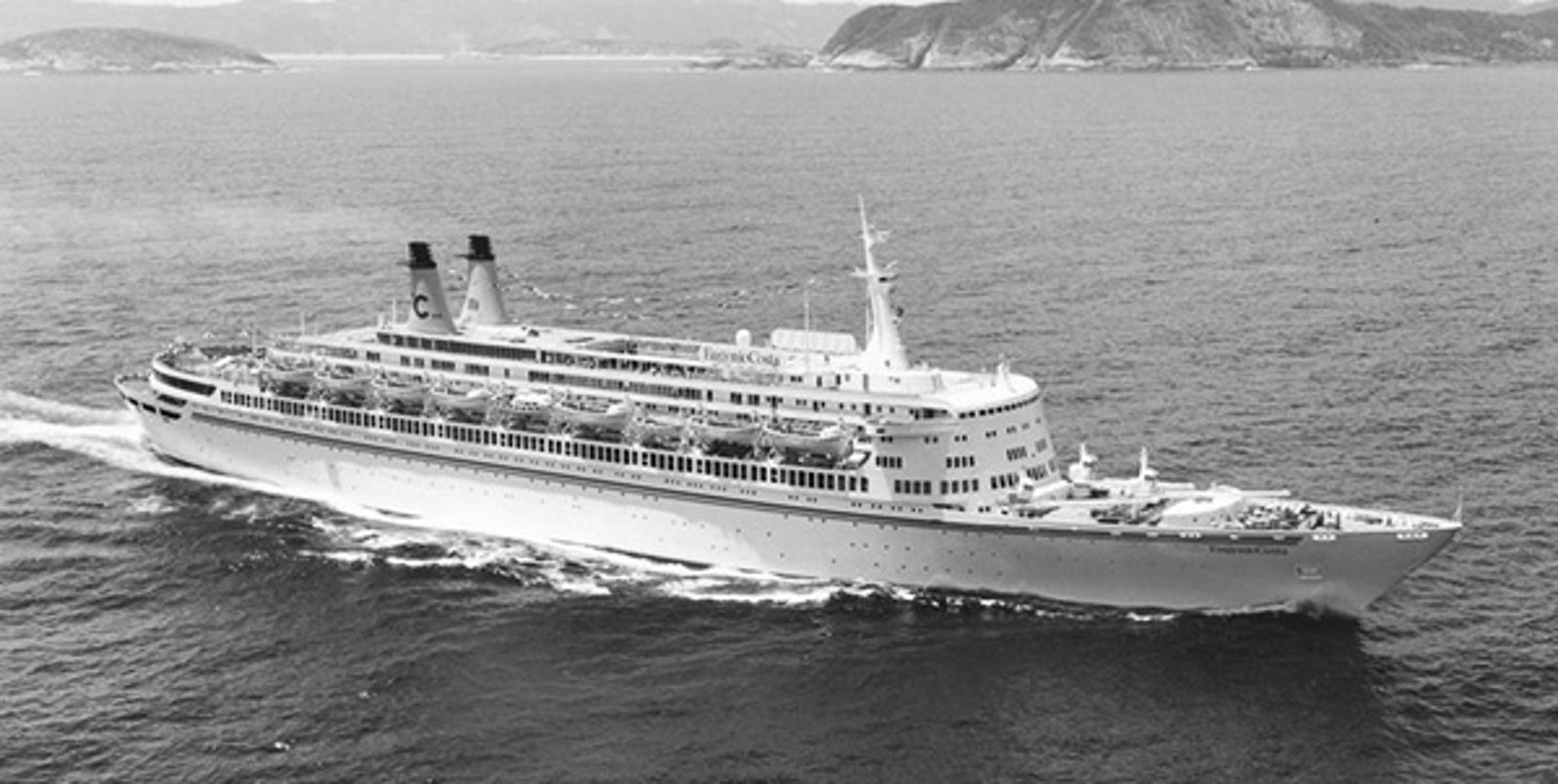 Fincantieri | About Us - Historic Ships: Black and white photo of a large cruise ship sailing on calm waters, multiple decks and two funnels with a visible 'C' logo, hilly background and serene marine setting, emphasizing the ship's scale and grandeur.