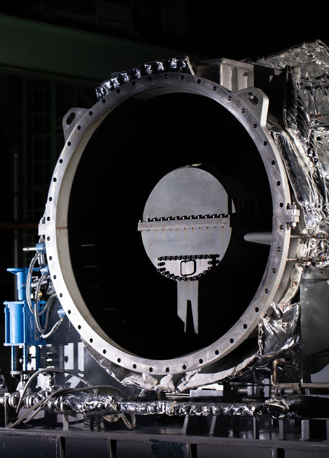 Fincantieri | Group - Innovation - Naval Ecosystem of the Future - Propulsion Systems and Clean Fuels: Large industrial machinery with a central circular component, factory environment with metallic and mechanical elements, details of bolts, wiring, and reflective surfaces, contrasting dark background.