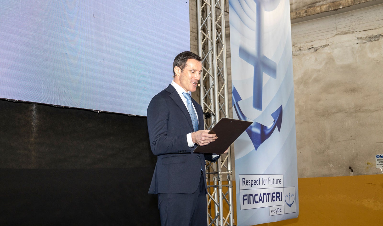 Suppliers and Partners – Fincantieri | Partnership: Man in formal attire on stage with a clipboard during a presentation, background with large screen and banner featuring the Fincantieri logo, “Respect for Future” text, nautical anchor and “Fincantieri S.p.A.” inscription, presence of industrial elements and stage lighting, 2nd Fincantieri Supplier Summit.