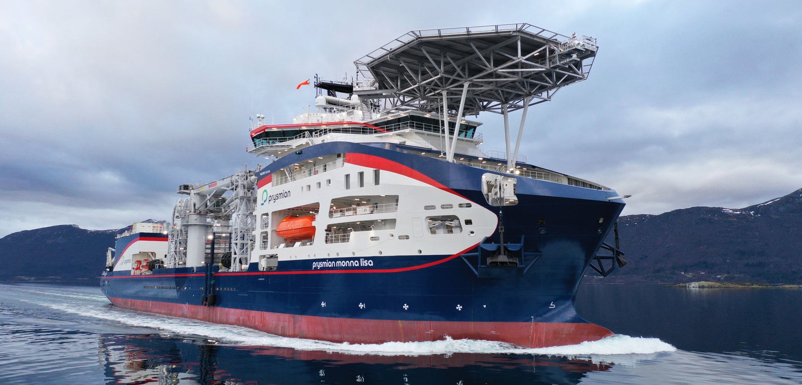 Prodotto - Offshore Vessel Normand Maximus: Vista laterale della nave operativa in navigazione, elisuperficie e scialuppe visibili, nome Normand Maximus chiaramente leggibile sullo scafo, sfondo montuoso e cielo nuvoloso.