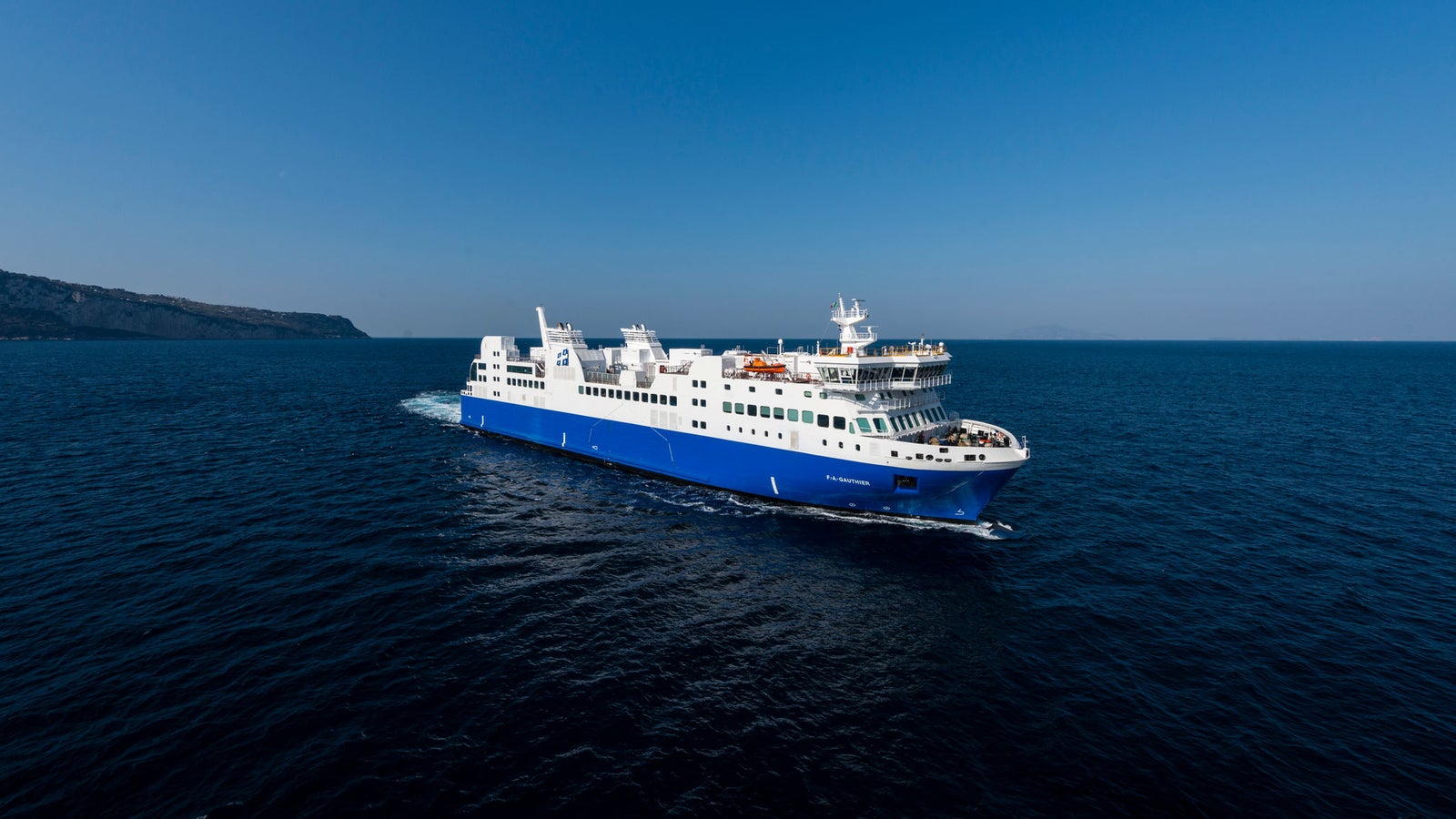 Fincantieri | Group - Business - Products - Ferries - Cruise FA Gauthier: Large blue and white ferry "FA Gauthier" in operation, multiple decks, visible lifeboats, sailing across deep blue sea, rocky coastline in background, clear sky.