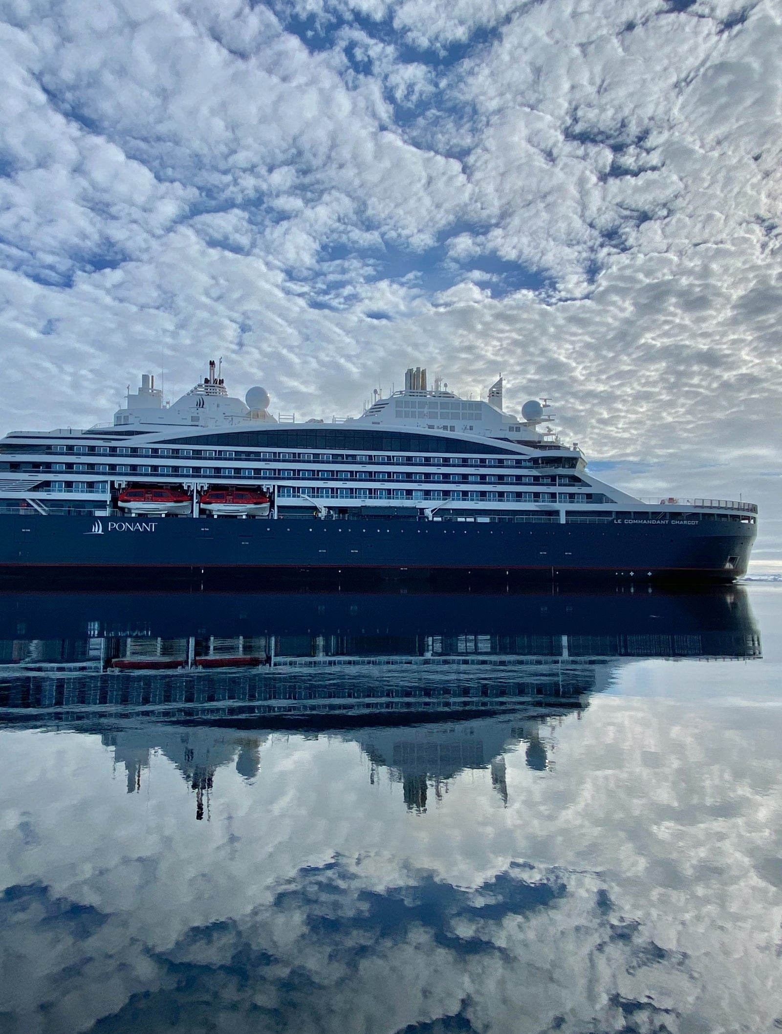 Fincantieri | Group - Business - Products - Cruise Ships - LE COMMANDANT CHARCOT: Large luxury cruise ship in operation reflected on still, mirror-like waters under partly cloudy sky, serene setting highlighting sleek design, bright sunlight through clouds.