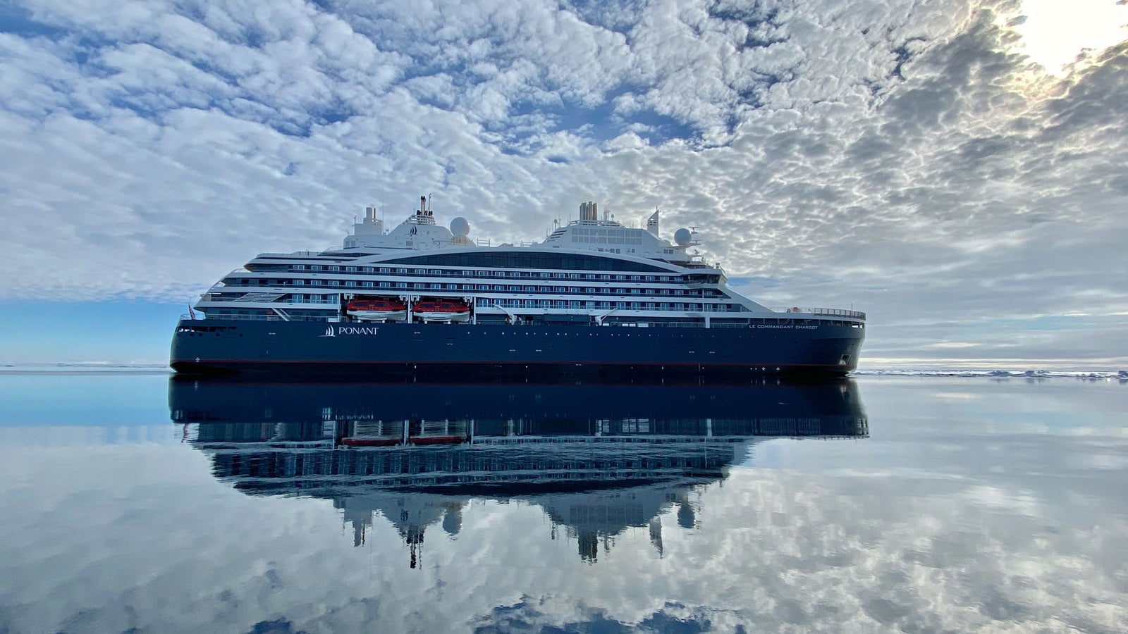 Fincantieri | Group - Business - Products - Cruise Ships - LE COMMANDANT CHARCOT: Large luxury cruise ship in operation reflected on still, mirror-like waters under partly cloudy sky, serene setting highlighting sleek design, bright sunlight through clouds.