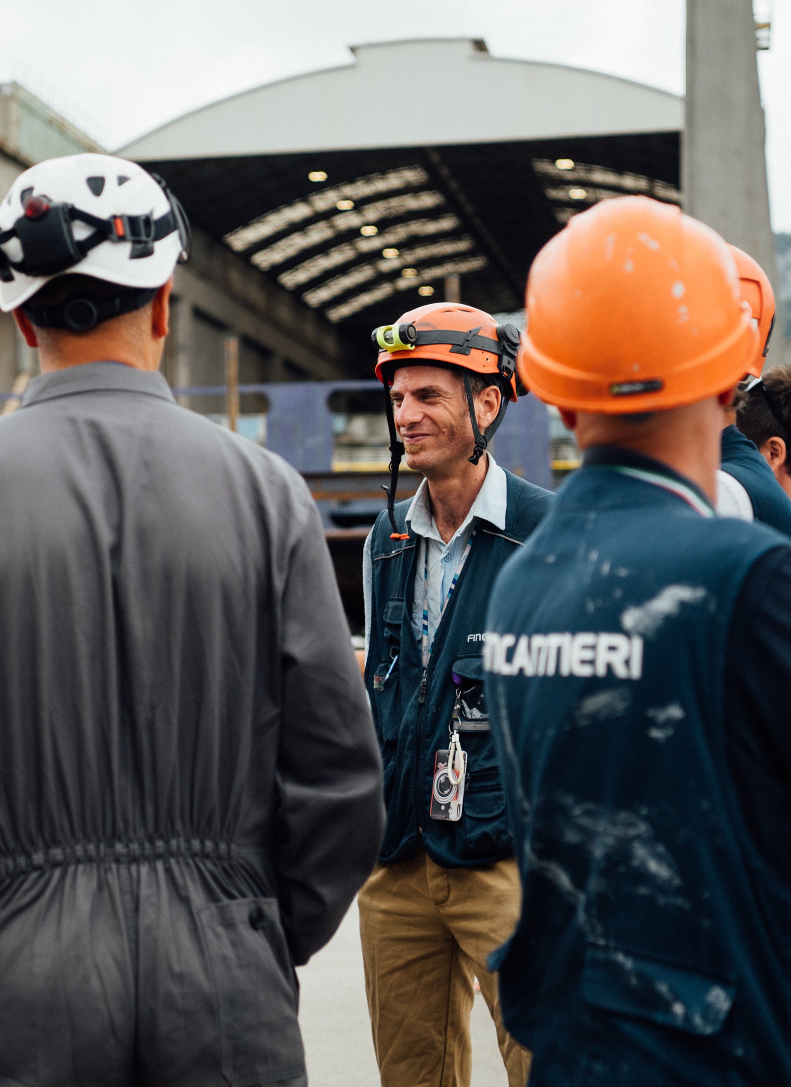 Fincantieri | Sostenibilità - Eccellenza industriale - Operatori cantiere: Gruppo di lavoratori con caschi, indumenti protettivi e headlamp in ambiente navale industriale, magazzino con tetto aperto e travi a vista sullo sfondo. Testo 'Fincantieri' visibile su un giubbotto. Atmosfera professionale e collaborativa.