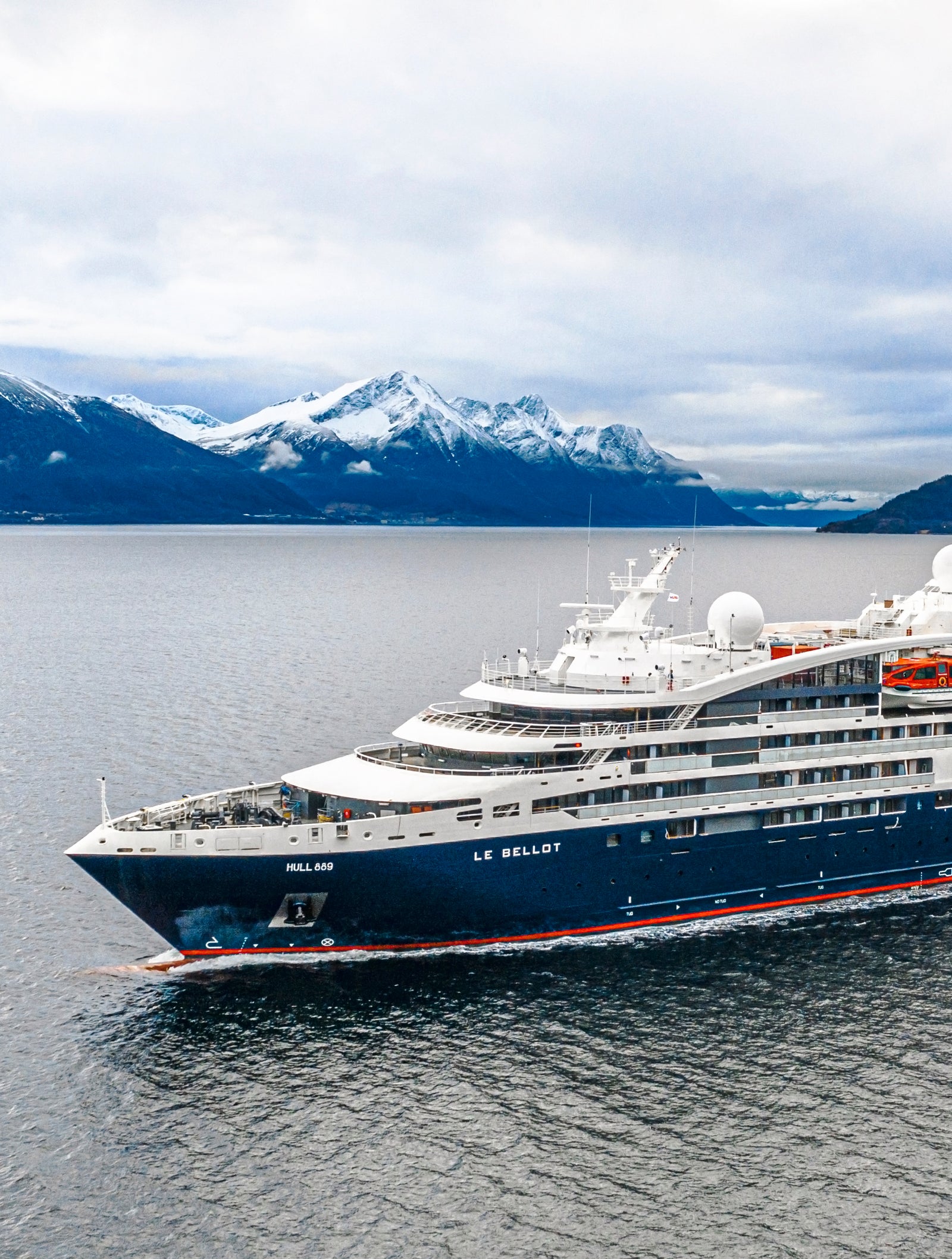 Fincantieri | Gruppo - Business - Prodotti - Navi da Crociera - Le Bellot: Vista laterale della nave da crociera Le Bellot operativa in navigazione all’interno di un fiordo, circondata da montagne innevate e acque calme che riflettono il paesaggio, scafo dal profilo lineare e cupole satellitari visibili sul ponte superiore.