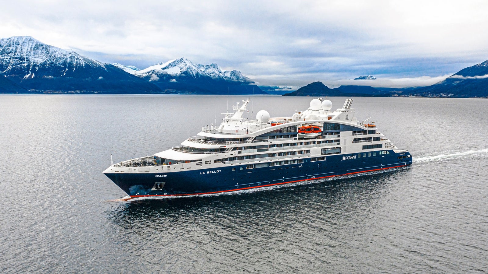  Fincantieri | Gruppo - Business - Prodotti - Navi da Crociera - Le Bellot: Vista laterale della nave da crociera Le Bellot operativa in navigazione all’interno di un fiordo, circondata da montagne innevate e acque calme che riflettono il paesaggio, scafo dal profilo lineare e cupole satellitari visibili sul ponte superiore.