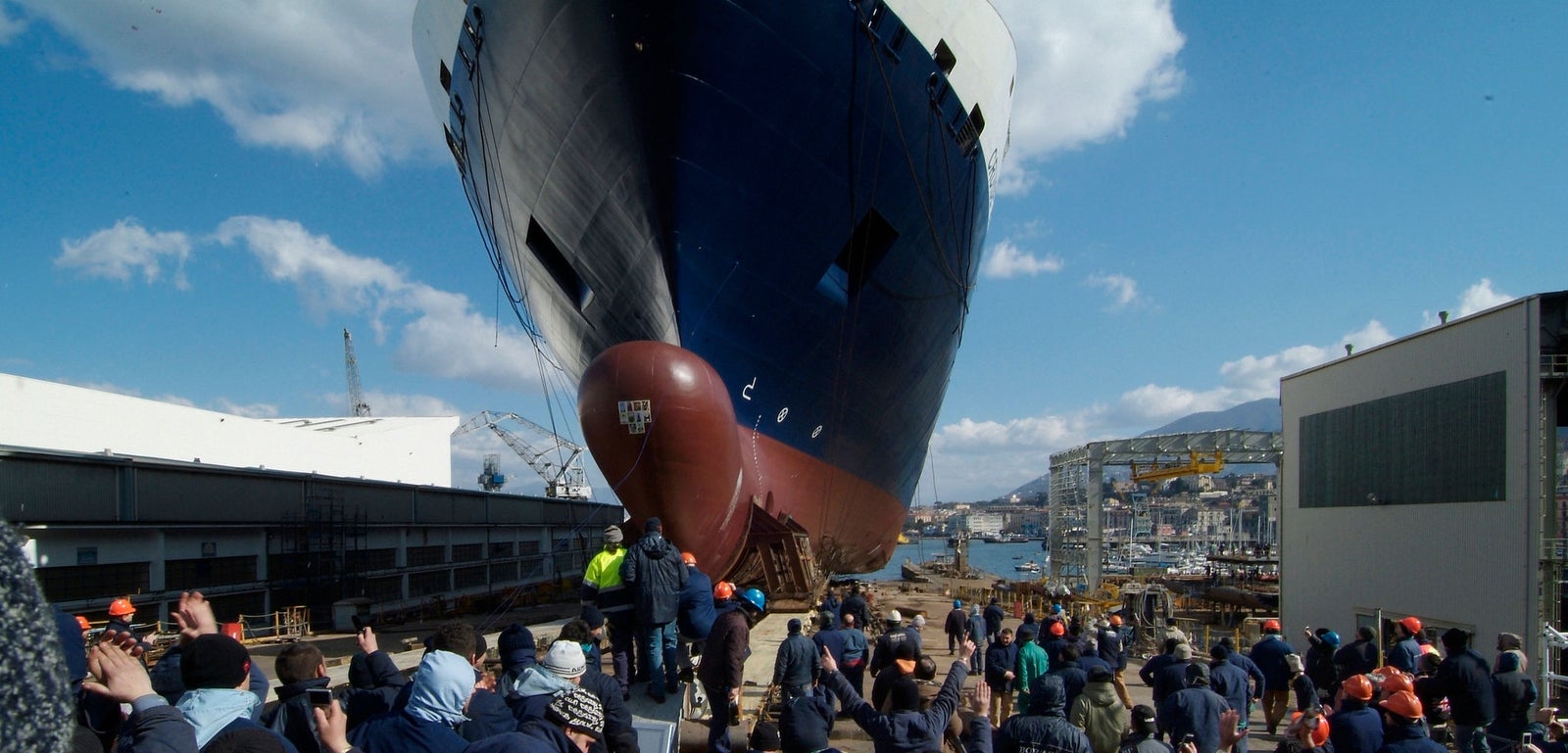 Fincantieri | Group - Business - Products - Ferries - Cruise Barcellona: Massive ship being launched at shipyard, surrounded by crowd of onlookers; industrial structures and clear blue sky; people dressed warmly, some in helmets and safety gear, indicating a cold day; ship's hull with visible markings enhances the maritime theme.