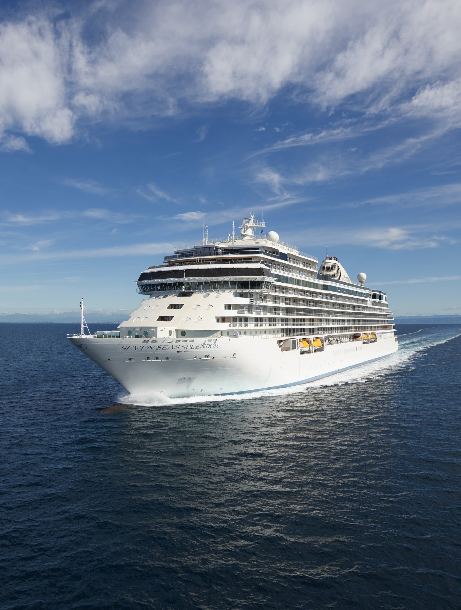 Fincantieri | Group - Business - Products - Cruise Ships - Seven Seas Splendor: Front view of large modern operational cruise ship with multiple decks and visible lifeboats, sailing on calm waters under clear sky, visible wake and sunlight reflecting on ocean, distant horizon completes serene maritime scene.