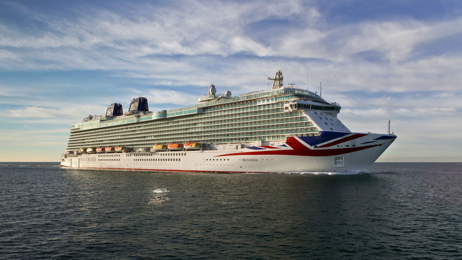 Fincantieri | Group - Business - Products - Cruise Ships - Britannia: View of Britannia cruise ship in operation navigating open sea, hull with red, white and blue Union Jack design on bow, visible lifeboats.