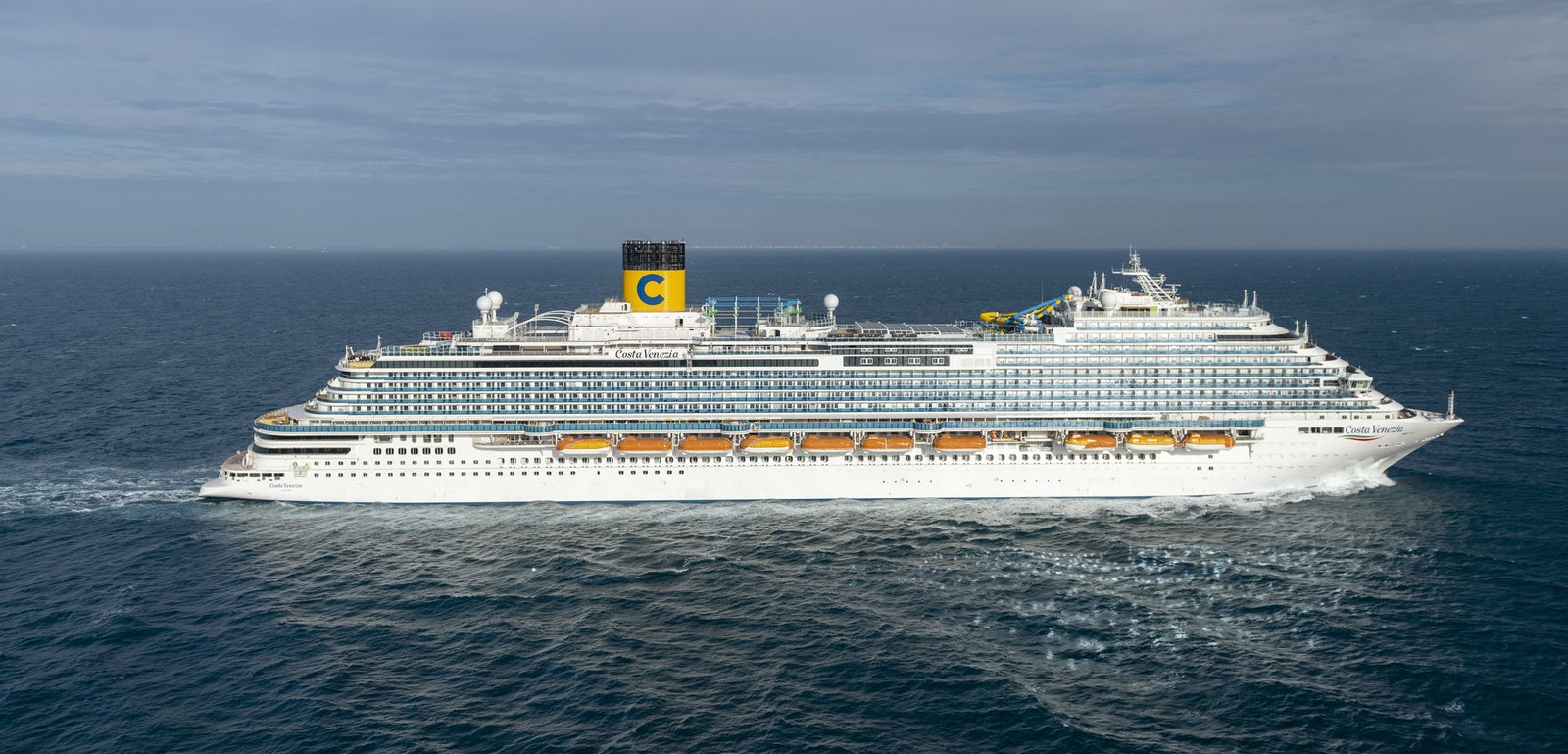 Fincantieri | Group - Business - Products - Cruise Ships - Costa Venezia: Large operational cruise ship with multiple decks, lifeboats and yellow funnel with 'C' logo clearly visible, sailing on calm waters under partly cloudy sky, imposing design and focus on leisure travel and maritime exploration.