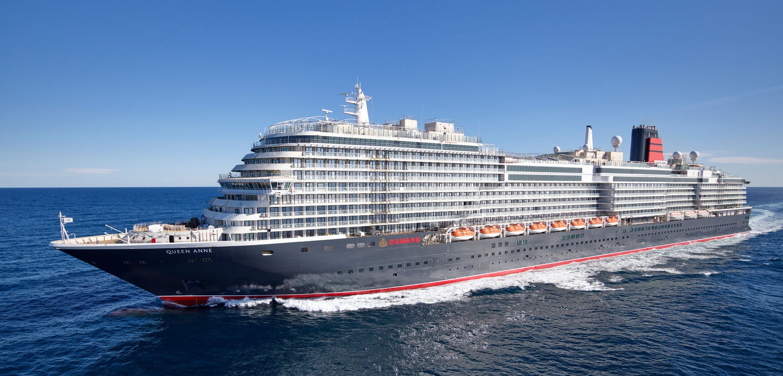 Fincantieri | Group - Business - Products - Cruise Ships - Queen Anne: Side view of Queen Anne cruise ship in operation, navigating open sea, linear profile hull, multiple passenger decks, name "Queen Anne" visible on hull.