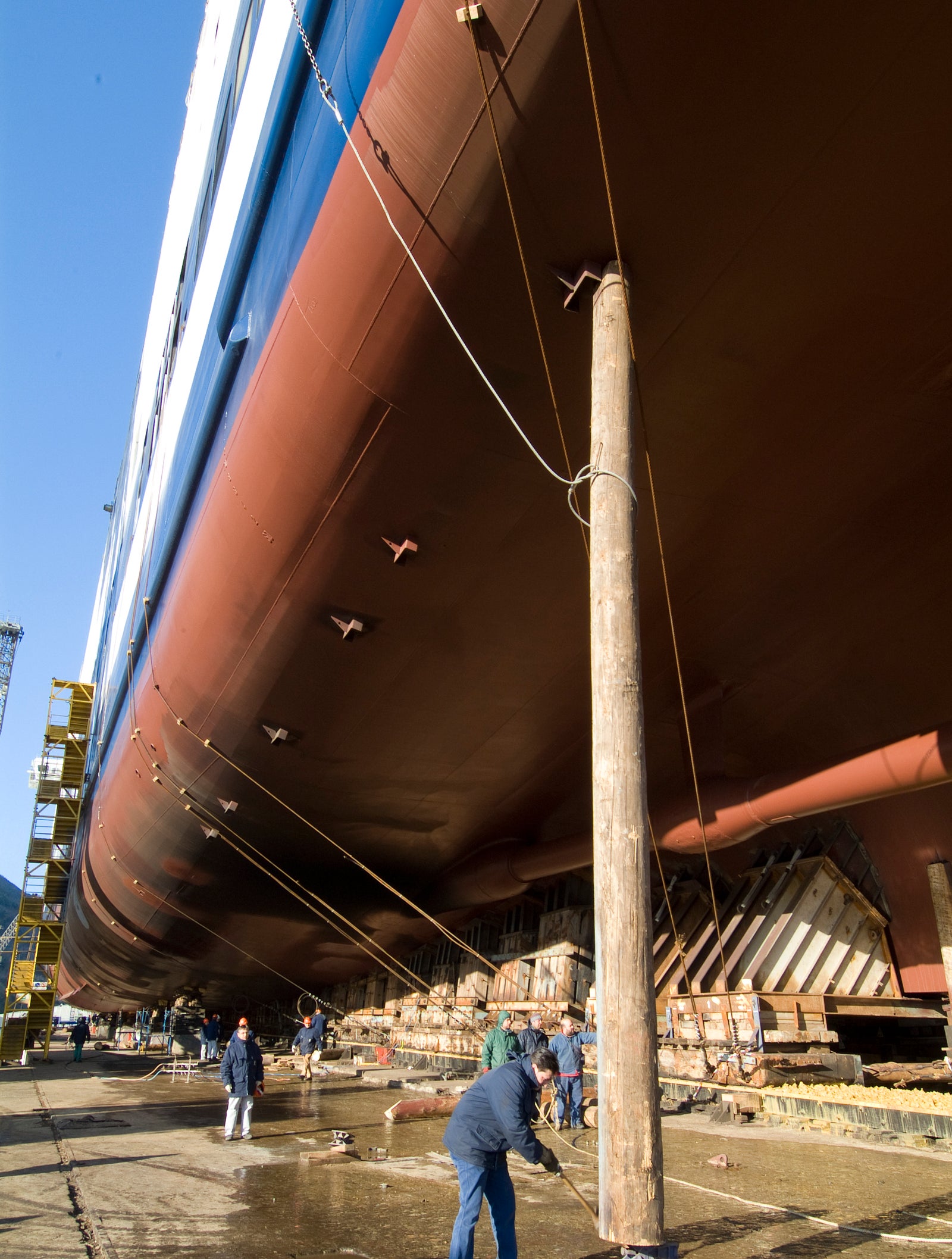  Fincantieri | Group - Business - Products - Ferries - Cruise Barcellona: Group of workers repairing large ship in dry dock setting; massive hull dominates the scene with support beams and scaffolding visible; workers engaged in maintenance activities under clear blue skies, emphasizing industrial and maritime environment.