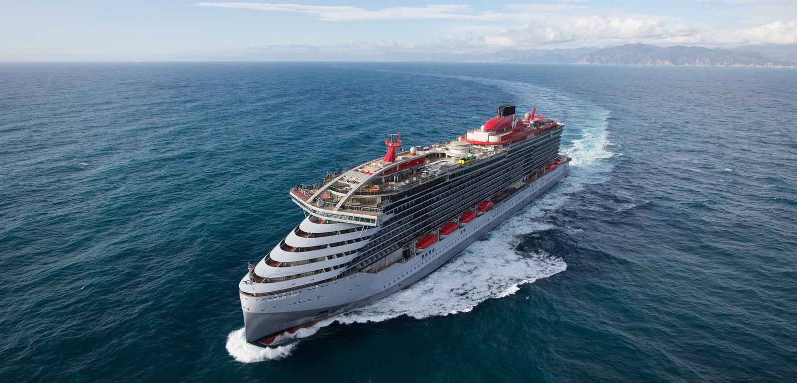 Fincantieri | Group - Business - Products - Cruise Ships - Scarlet Lady: Modern operational cruise ship with multiple decks, red lifeboats and distinctive red funnel, sailing on calm waters under partly cloudy sky, coastline visible in background.