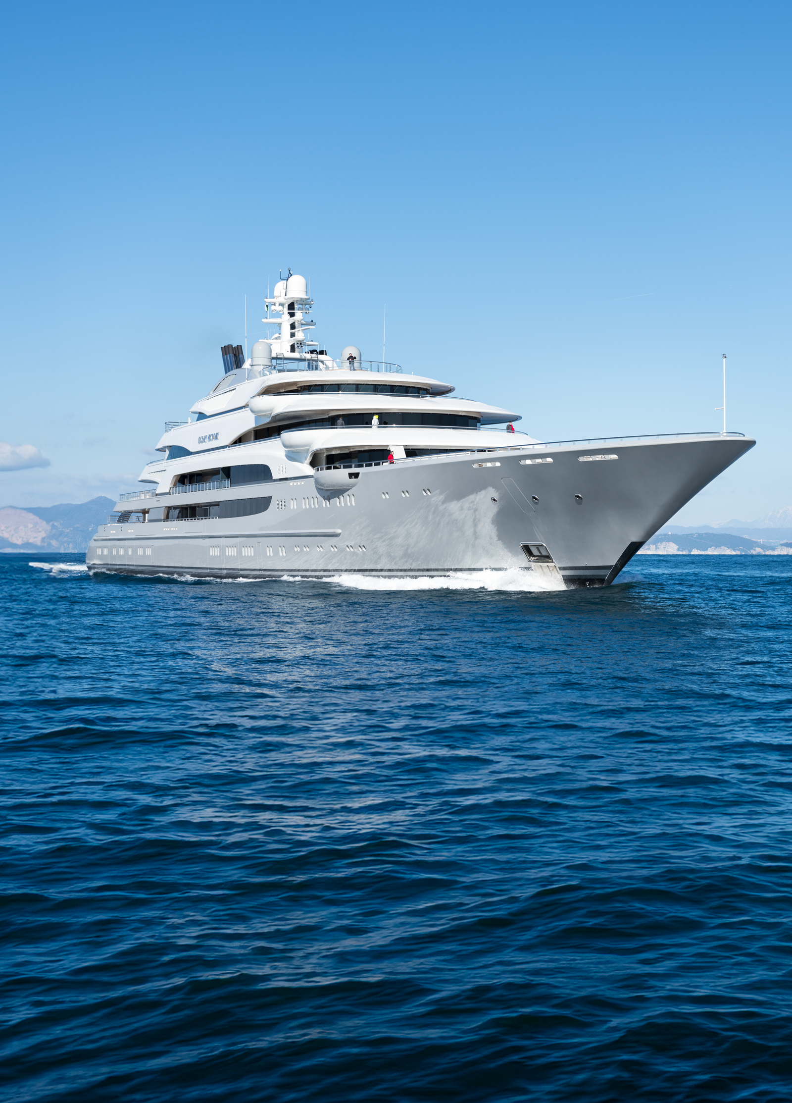 Fincantieri | Services - Yacht Repair and Conversion: Side view of a modern luxury yacht sailing on calm blue waters, multi-deck design with white and grey accents, background with distant mountains, elegant maritime setting.