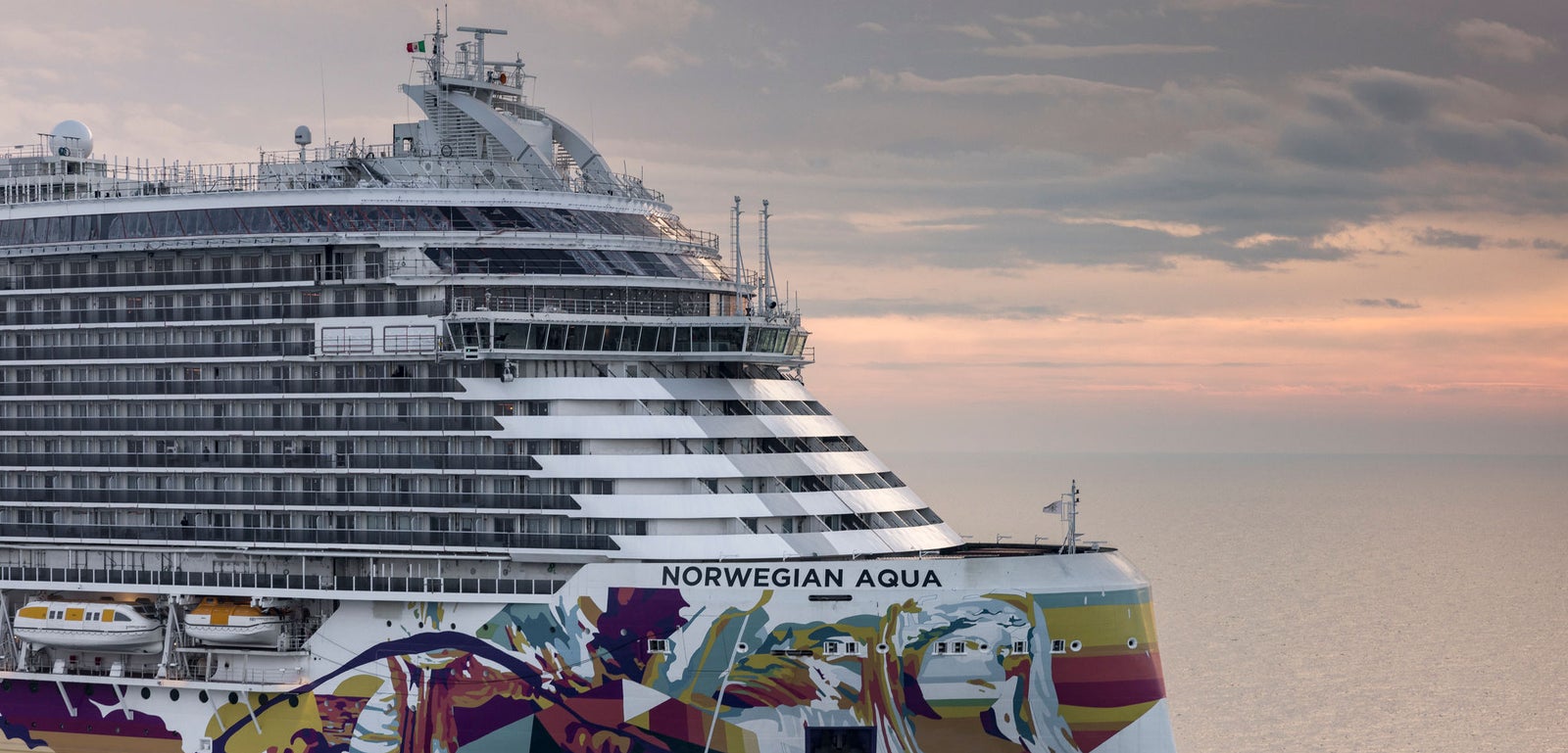 Fincantieri | Group - Business - Products - Cruise Ships - Norwegian Aqua: Large operational cruise ship with vibrant and colorful artwork on the hull, side view, sailing at sunset.