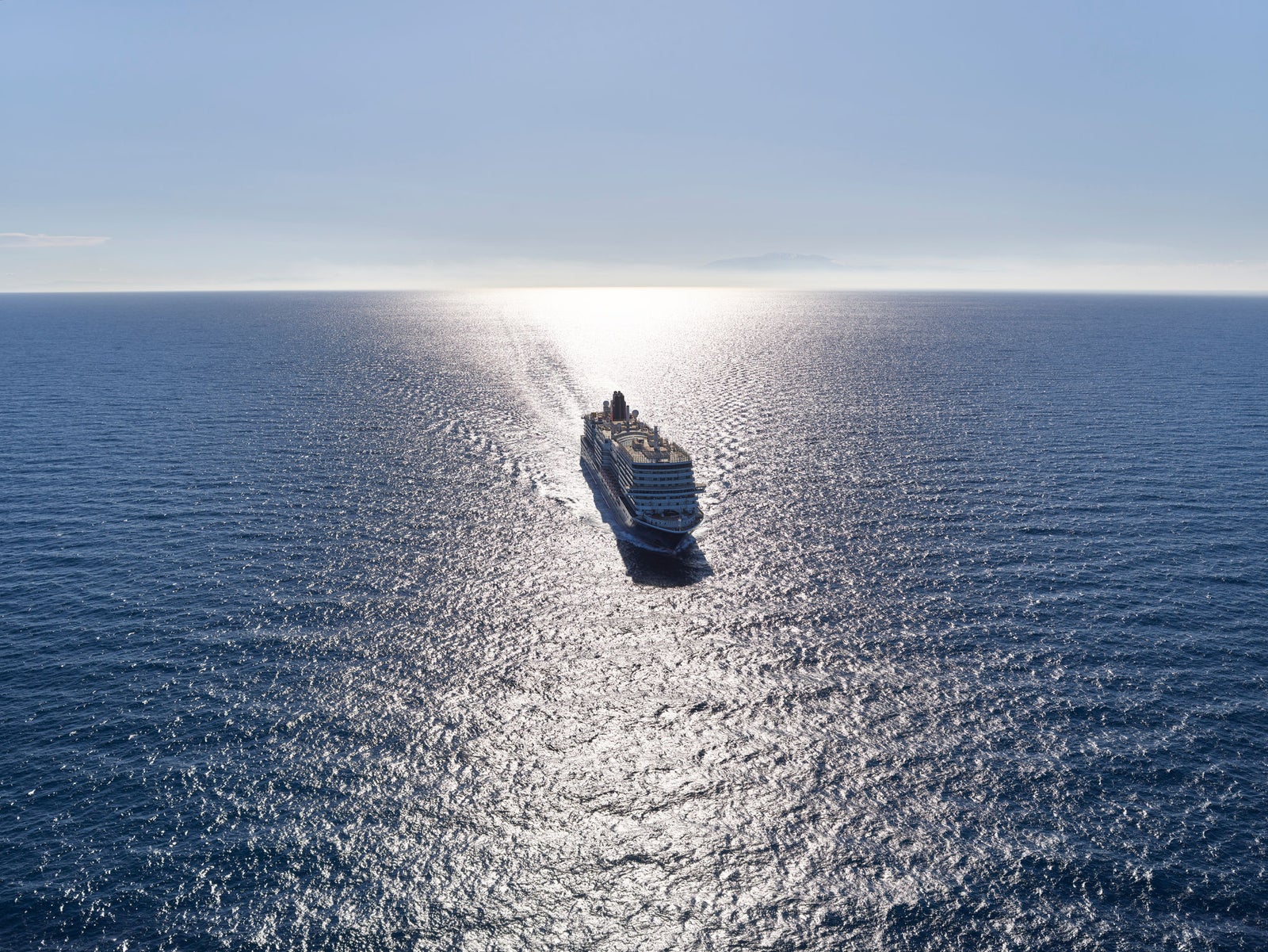 Fincantieri | Group - Business - Products - Cruise Ships - Queen Anne: Large cruise ship in operation, centered in frame, traveling across vast calm ocean, sunlight reflecting off water, clear blue sky.