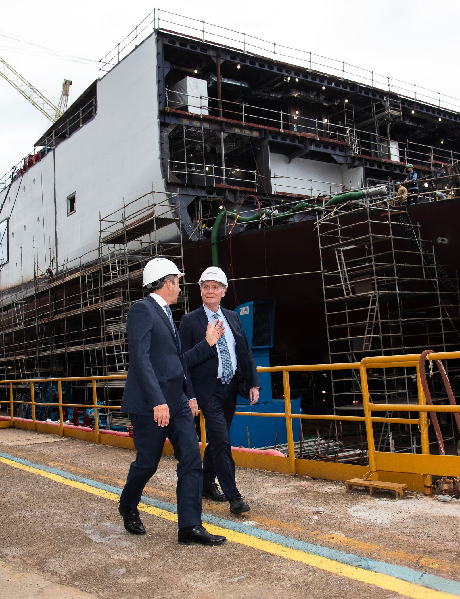 Fincantieri | Group - Sustainability - Responsible Culture - Sustainability Governance: Two men in formal attire, one wearing a white helmet, walk in a shipyard with a ship under construction, visible scaffolding and industrial equipment; environment of professional inspection or evaluation of the shipbuilding process.