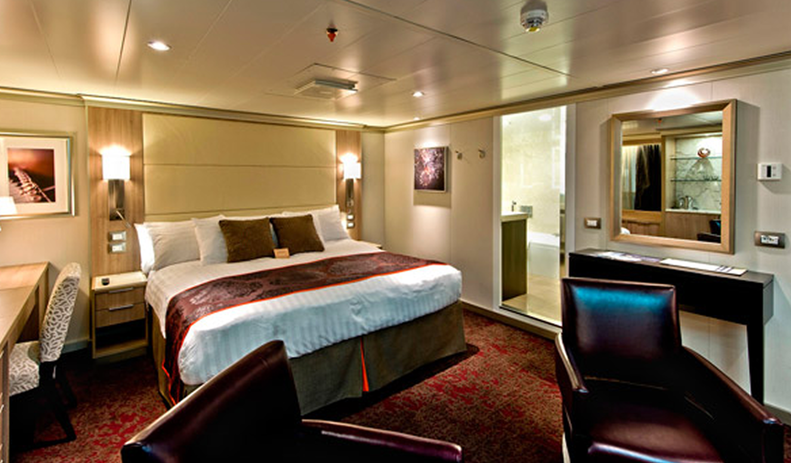 Fincantieri | About Us - Heritage and Future: Spacious, elegant hotel room with a large bed and soft linens, desk and relaxation area, warm lighting, glass-walled bathroom and modern furnishings, red carpet and neutral walls, welcoming atmosphere.