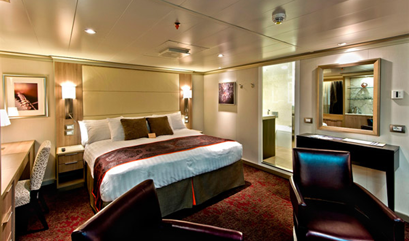 Fincantieri | Chi siamo - Eredità e Futuro: Ampia camera d’hotel elegante con letto grande e biancheria soffice, scrivania e zona relax, illuminazione calda, bagno vetrato e arredi moderni, moquette rossa e pareti neutre, atmosfera accogliente.