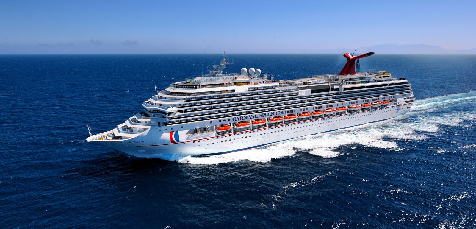 Fincantieri | Group - Business - Products - Cruise Ships - Carnival Splendor: Large operational cruise ship gliding on calm deep blue waters under clear sky, multiple decks, lifeboats, distinctive red and blue funnel, scenic background with distant mountains and tranquil horizon emphasizing the serenity of the journey.