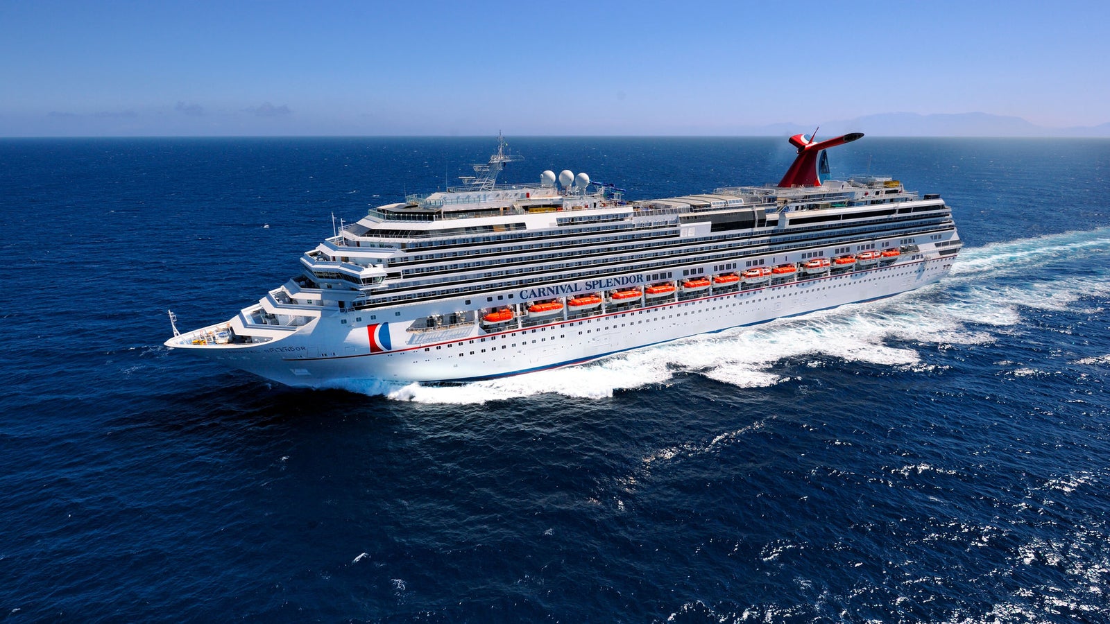 Fincantieri | Group - Business - Products - Cruise Ships - Carnival Splendor: Large operational cruise ship gliding on calm deep blue waters under clear sky, multiple decks, lifeboats, distinctive red and blue funnel, scenic background with distant mountains and tranquil horizon emphasizing the serenity of the journey.