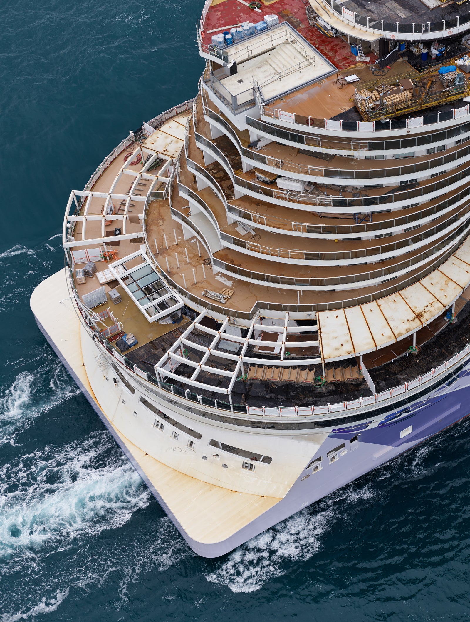  Fincantieri | Gruppo - Business - Prodotti - Navi da Crociera - Norwegian Viva: Vista aerea di grande nave da crociera in costruzione su acque aperte, molteplici ponti e dettagli complessi, materiali edili e sezioni incomplete visibili, scafo blu e bianco con motivo ondulato, oceano sullo sfondo.