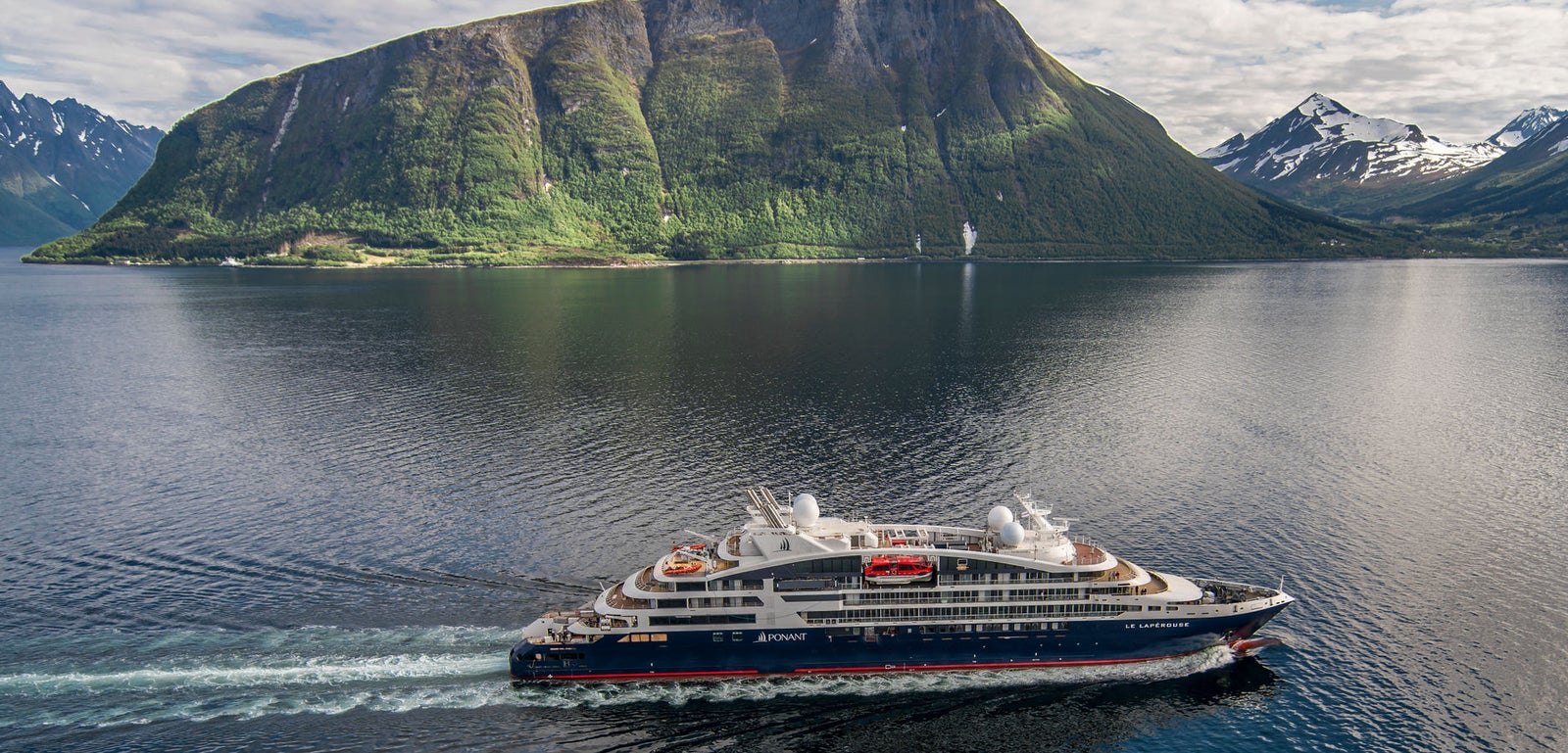  Fincantieri | Gruppo - Business - Prodotti - Navi da Crociera - Le Lapérouse: Vista laterale della nave Le Lapérouse operativa in navigazione in un fiordo, circondata da montagne verdi e picchi innevati riflessi nell’acqua calma, scafo bianco con dettagli rossi visibili.