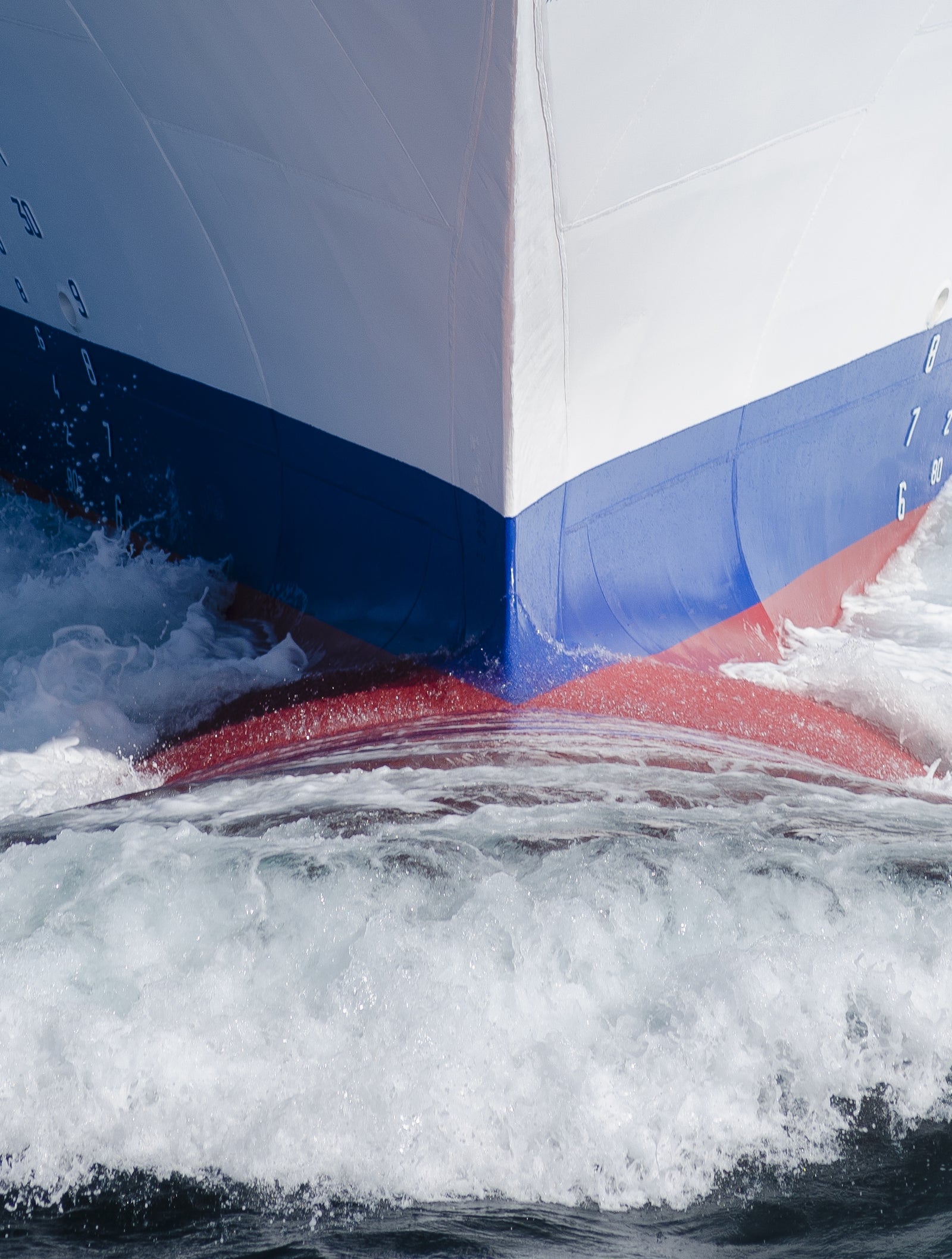 Fincantieri | Group - Business - Products - Cruise Ships - Costa Fascinosa: Close-up of Costa Fascinosa cruise ship bow in operation cutting through water, hull with red and blue details, visible spray.