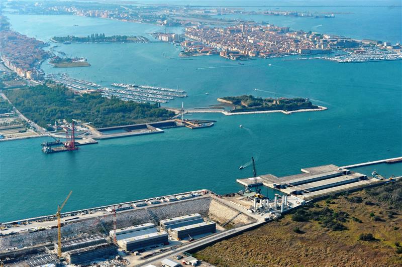 Fincantieri | Group - Subsidiaries and Affiliates - Fincantieri Opere Marittime: Expansive aerial view of coastal construction site near large body of water, cranes, docks, and infrastructure development visible; background includes cityscape and marina surrounded by waterways, highlighting industrial activity and urban proximity.